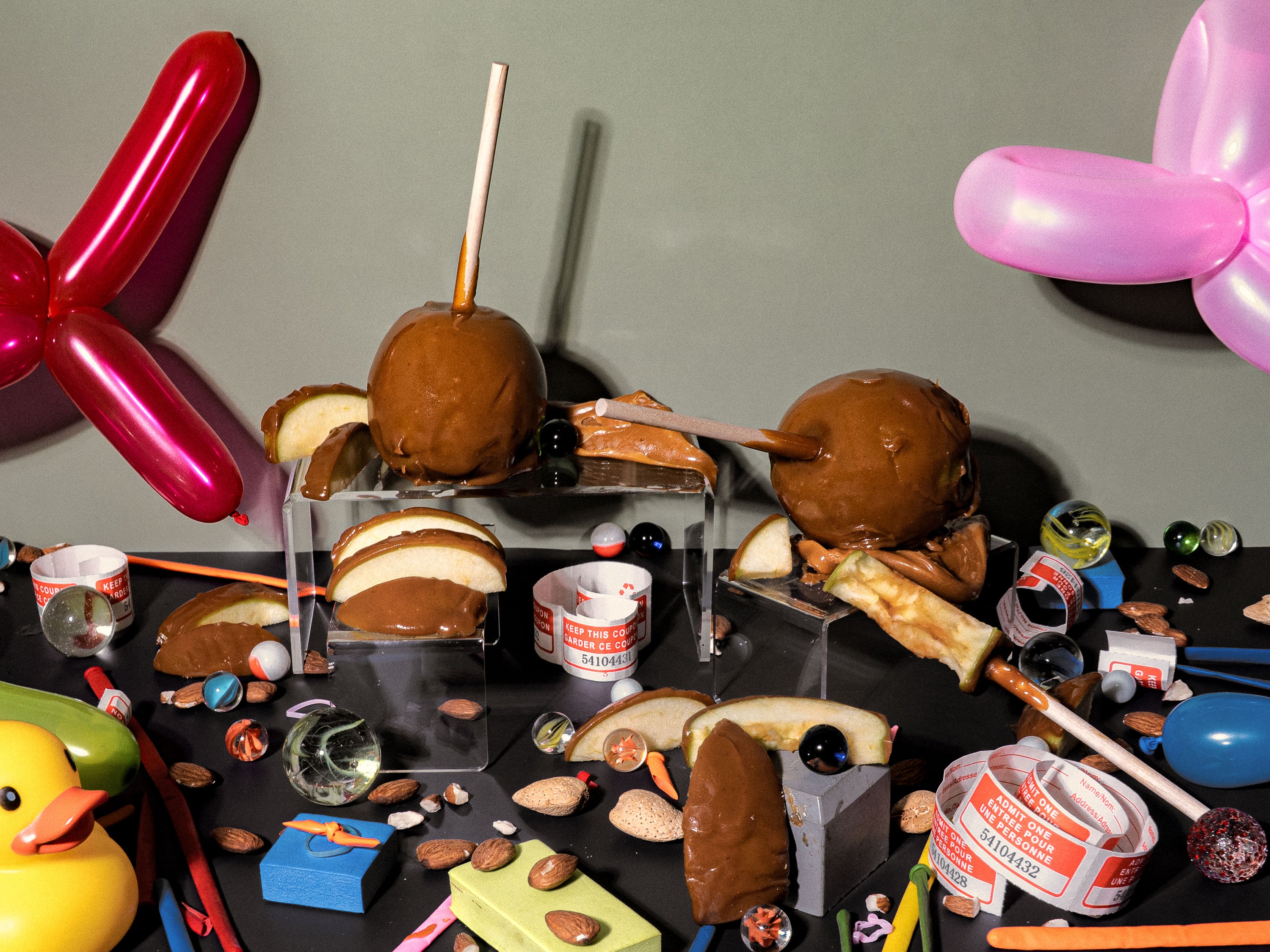 Whimsical flat lay, for a fragrance oil, featuring caramel apples, sliced green apples, marbles, almonds, carnival tickets, and colourful balloon animals. The nostalgic setup evokes the playful chaos of a county fair after dark, with sticky treats, scattered toys, and candy-coloured memories.