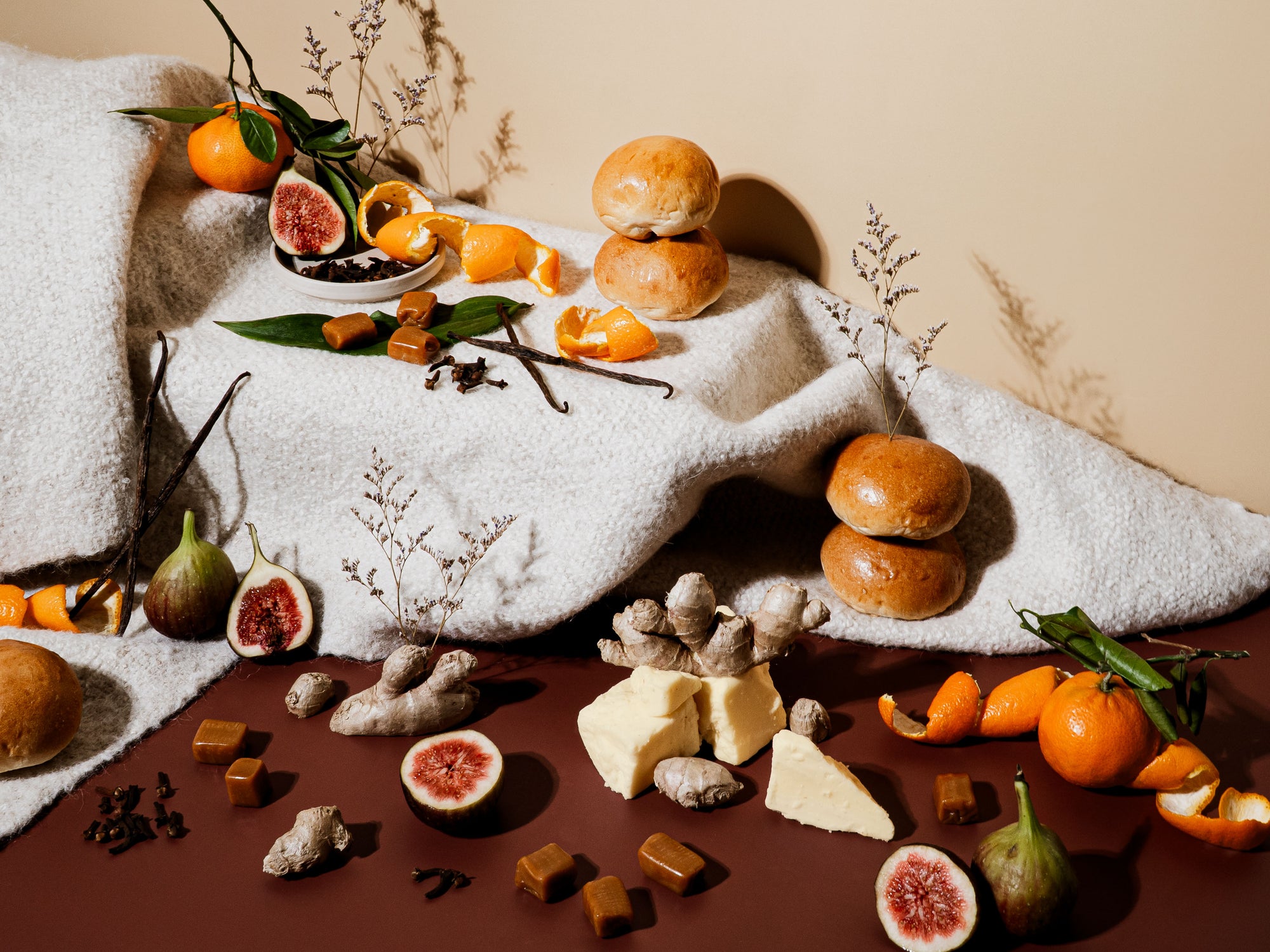 A cozy still life set on a warm brown surface and cream blanket, featuring figs, ginger root, tangerines with peels, clove, vanilla beans, soft rolls, and caramel pieces. The scene is rich with texture and warmth, evoking baked goods, mulled spices, and winter indulgence with a soft glow of comfort.