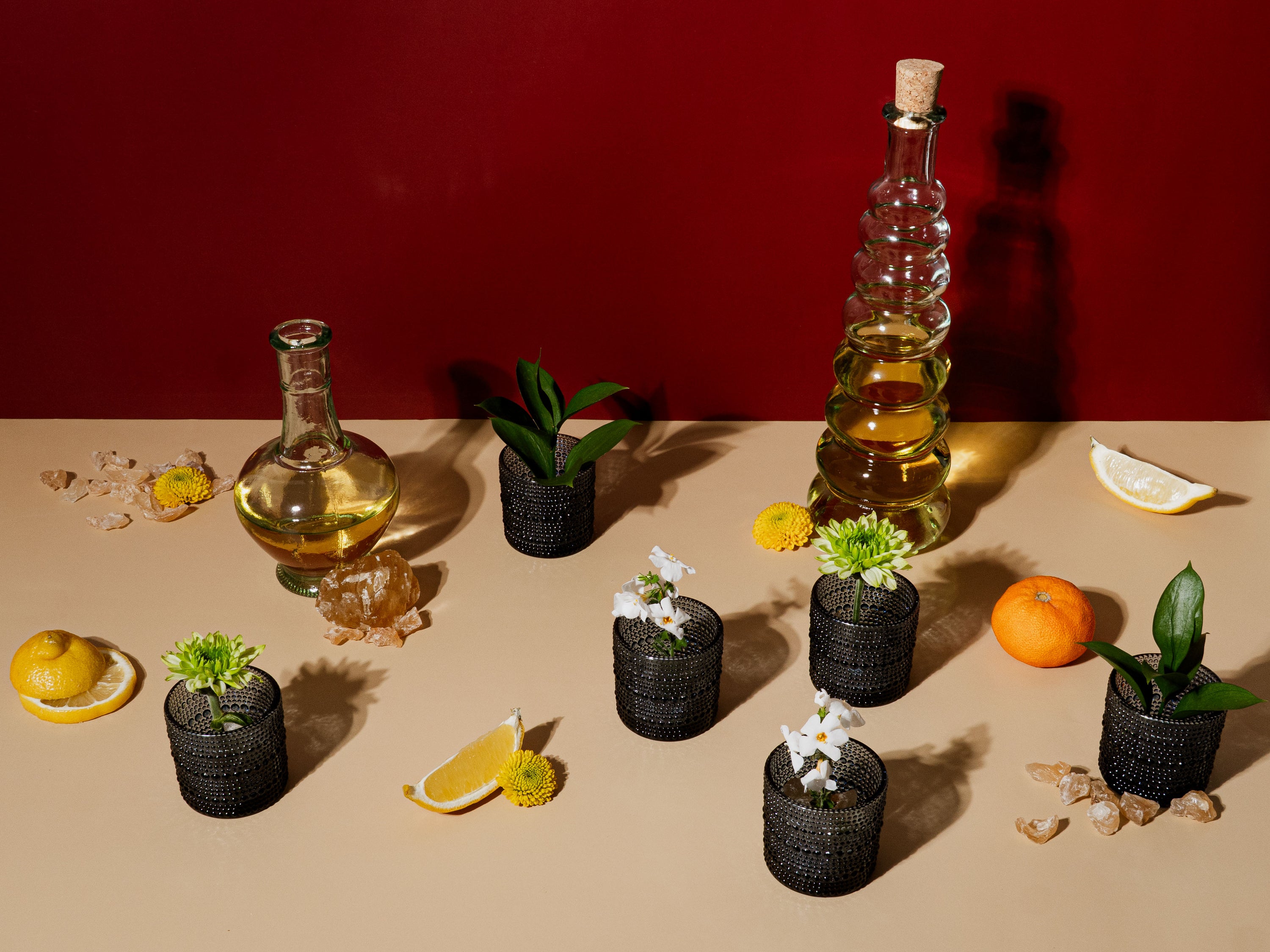 A still life on a beige surface with a deep red backdrop, featuring glass bottles filled with golden oil, citrus fruits, lemon wedges, fresh greenery, and small black textured vases holding white and yellow flowers. Amber resin pieces and slices of lemon and orange are scattered around, evoking a warm, radiant mood inspired by ritual and light.