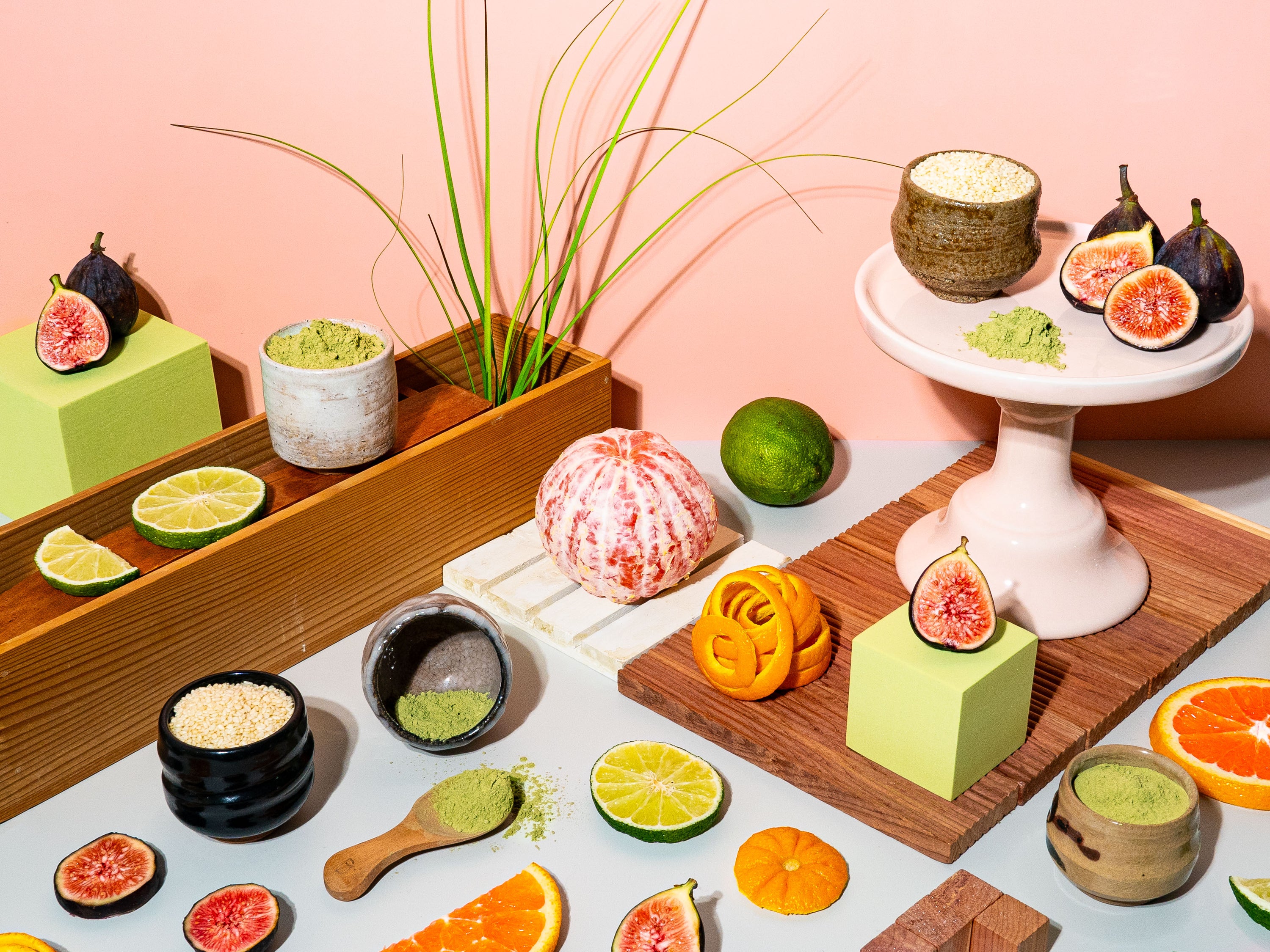 A vibrant, carefully arranged still life featuring fresh figs, citrus slices, matcha powder, and grains against a warm peach toned backdrop. Earthy matcha and bright limes sit alongside rich figs and fragrant orange peels, creating a scene that feels both natural and curated. Ceramic bowls and wooden surfaces add texture, evoking the balance of freshness and warmth. The composition is playful yet refined, celebrating the depth of scent and flavour in each element.