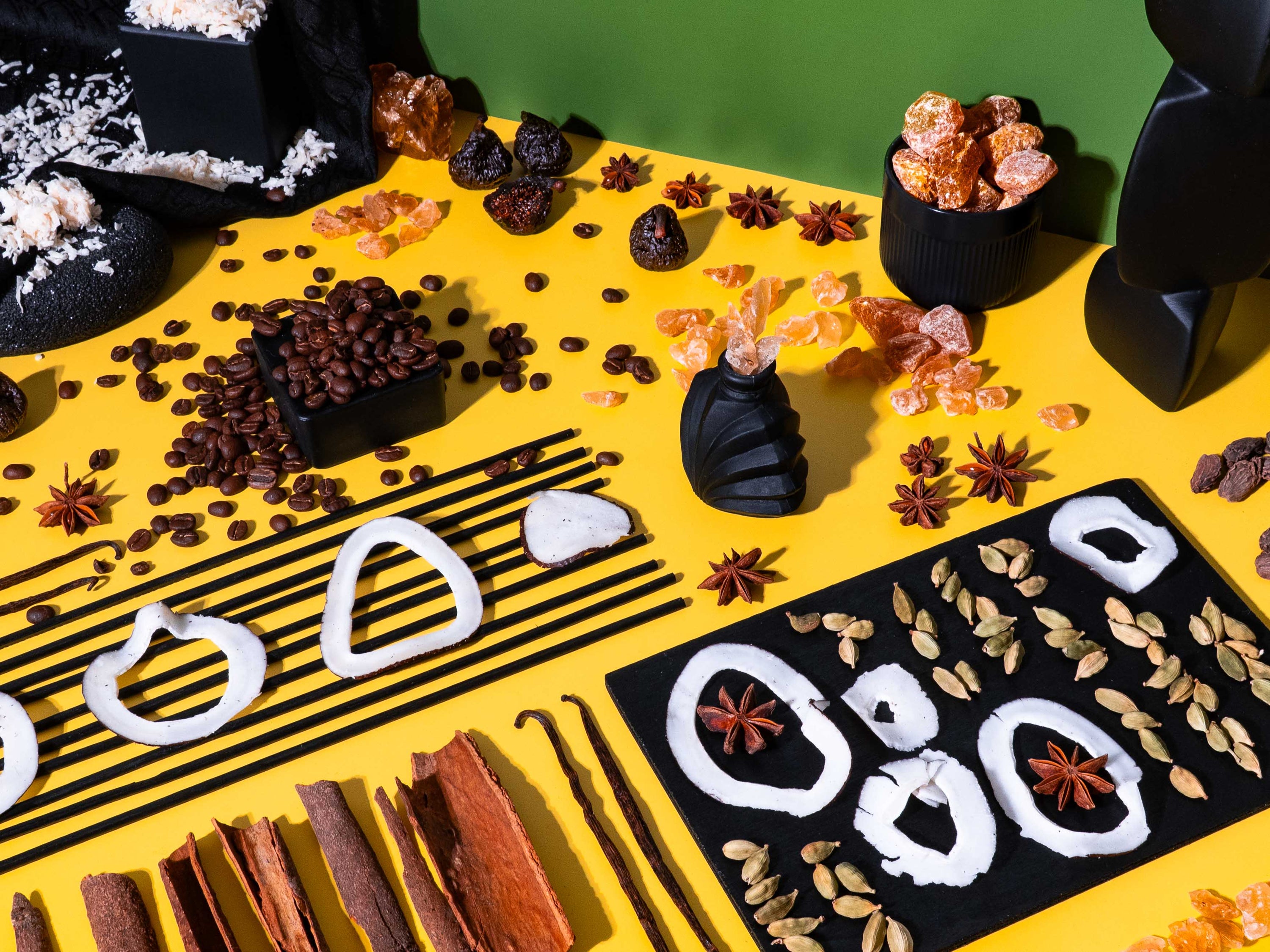 Flat lay of fragrance oil ingredients on a split yellow and green background featuring coffee beans, coconut rings, cardamom pods, cinnamon sticks, star anise, vanilla beans, dried figs, and rock sugar, arranged in a modern, graphic composition with black ceramic vessels and abstract shapes.