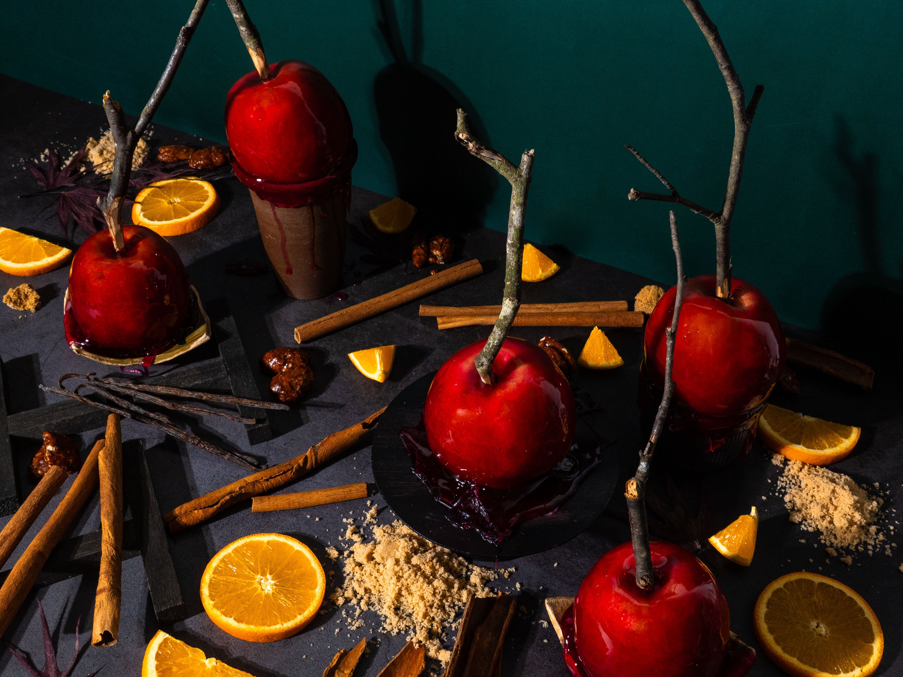 Cinematic flat lay for a fragrance oil, of candy apples on dark surface with cinnamon sticks, brown sugar, orange slices, and vanilla beans, styled with rustic twigs and deep shadows. Autumn carnival-inspired fragrance photography.