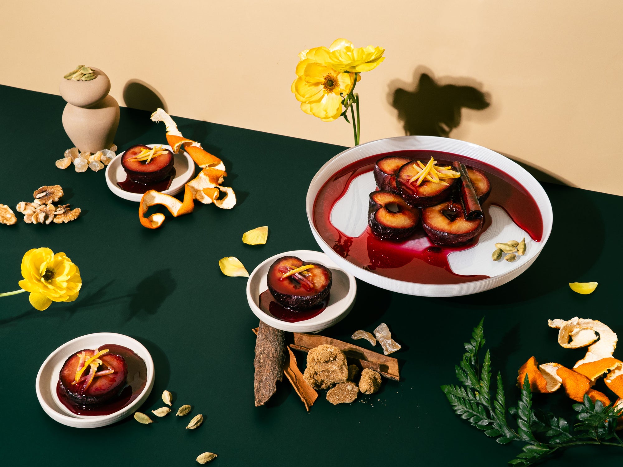 A dramatic still life on a dark green surface featuring halved plums glazed in red syrup, garnished with citrus peel and cardamom. The scene includes bowls of poached plums, cinnamon bark, orange peels, yellow flowers, brown sugar, walnuts, and greenery, evoking a rich, spiced dessert with a deep, sensual warmth.