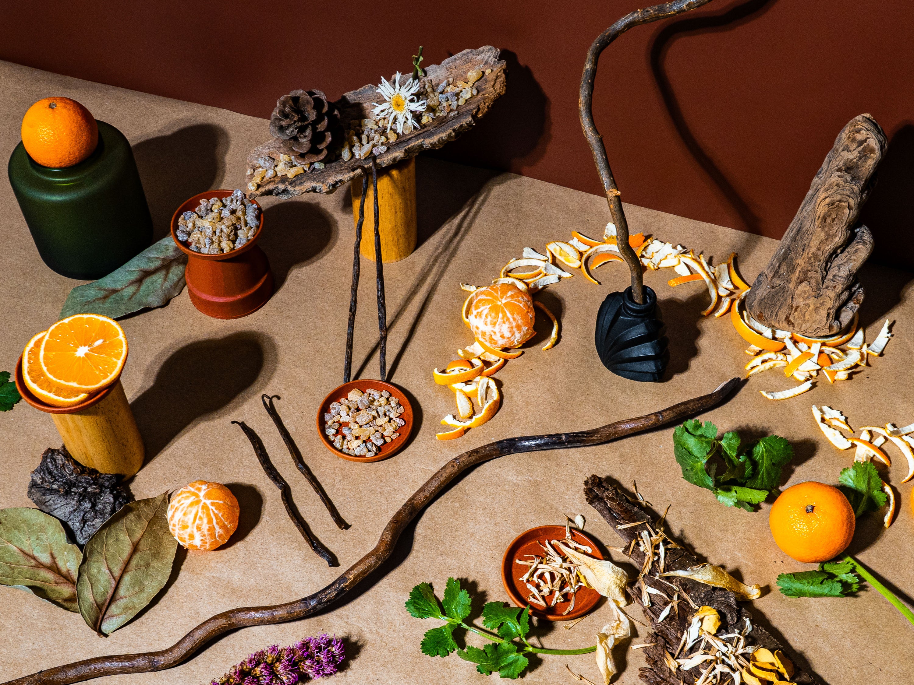 A visually rich flat lay composition featuring citrus elements, including whole and sliced oranges and tangerines, dried botanicals, vanilla pods, and fresh herbs arranged on a neutral-toned background. Wooden textures, pinecones, and resin pieces are artfully placed, accompanied by a black sculptural object and a curved branch, adding depth and contrast. Orange peels are scattered around, enhancing the natural, earthy aesthetic of the scene. The lighting casts soft shadows, emphasizing the details