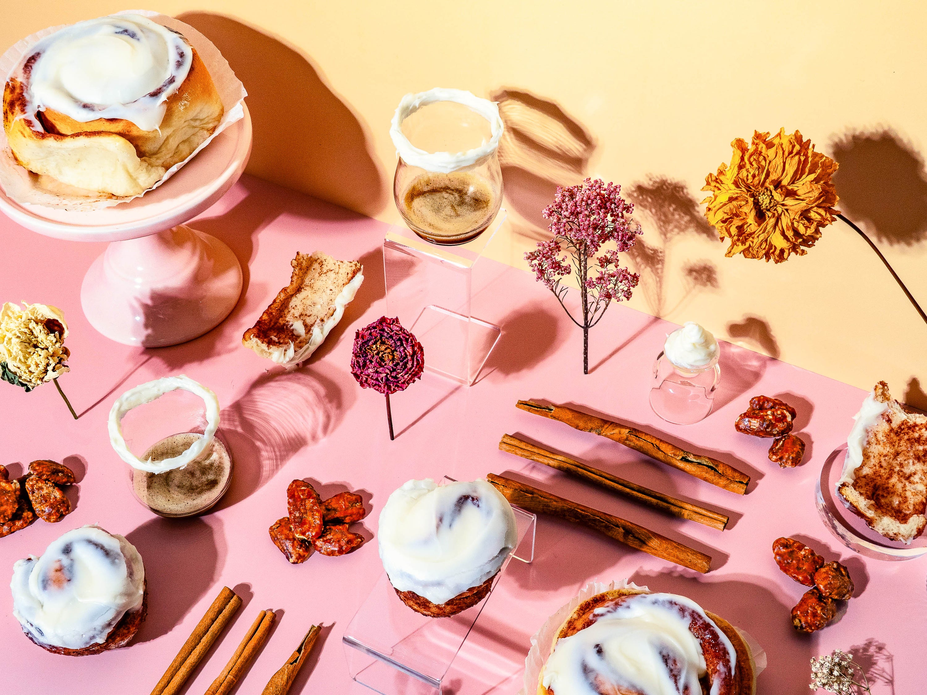 Aesthetic flat lay for a fragrance oil, featuring frosted cinnamon rolls, whole cinnamon sticks, whipped vanilla cream, dried flowers, caramelized nuts, and dessert crumbs arranged on a pink and peach background with modern glass accents and soft natural lighting.