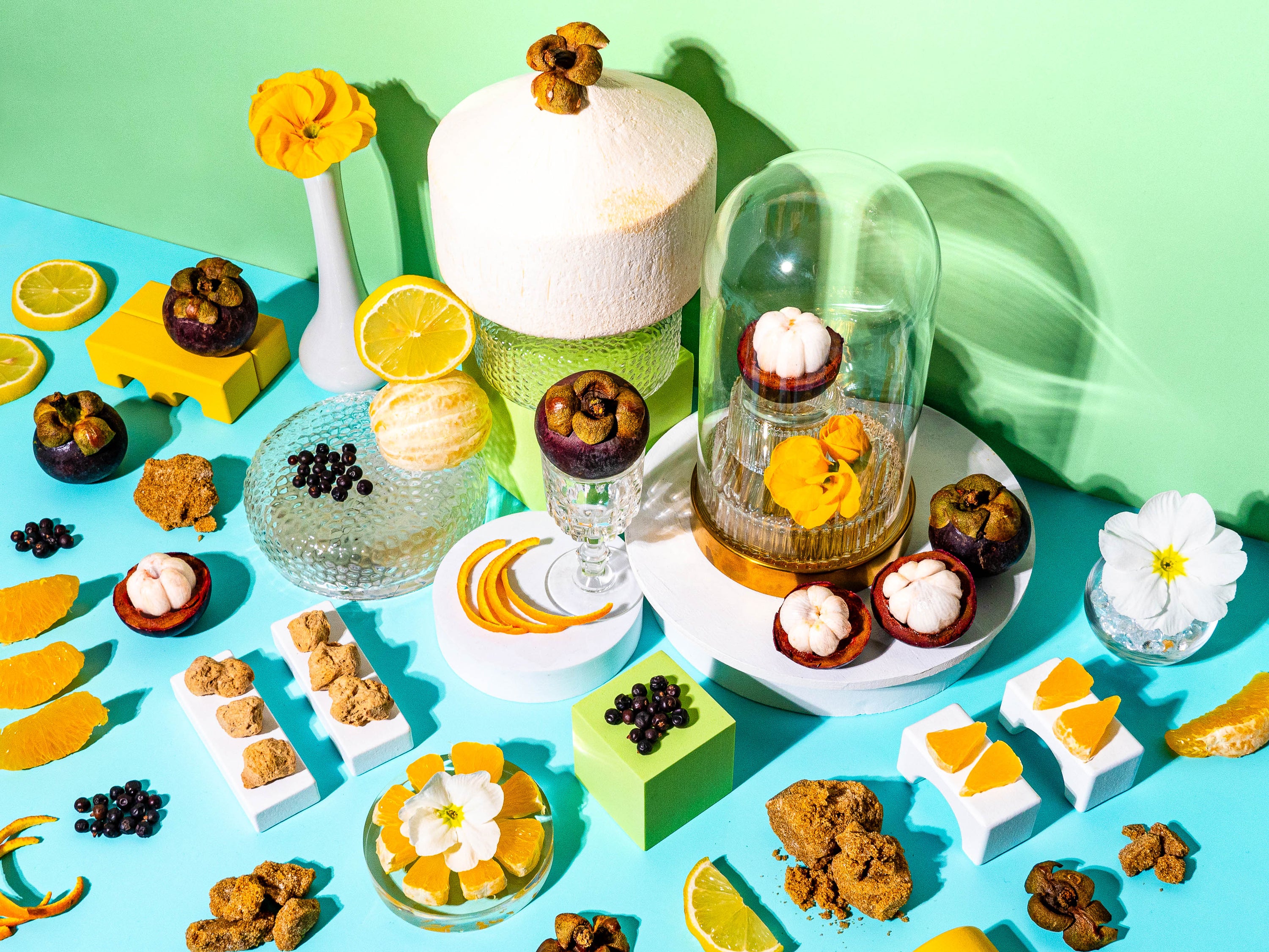 A vibrant flat-lay composition featuring tropical ingredients against a bright turquoise and green background. A whole coconut sits on a textured glass pedestal, surrounded by halved mangosteens, fresh citrus slices, black currants, and golden sugar-coated treats. A glass cloche encases a single mangosteen and delicate yellow flowers, adding an elegant touch. Scattered lemon slices, orange peels, and white blossoms enhance the summery, sunlit feel of the scene, evoking the a fresh, tropical fragrance.