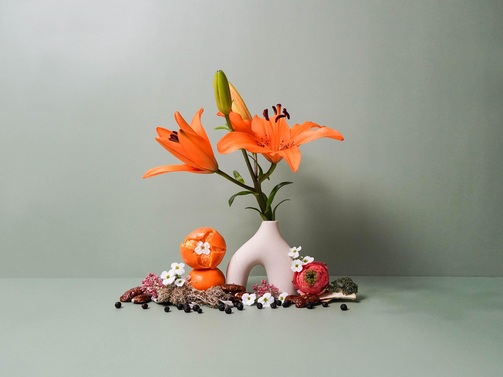 A minimalist still life composition featuring vibrant orange lilies in a modern white ceramic vase, surrounded by scattered botanicals and textures. The arrangement includes a halved orange, delicate white and pink flowers, green moss, dark juniper berries, and earthy spices. Set against a muted sage green background, the scene evokes a balance of bold colour and serene natural elements.