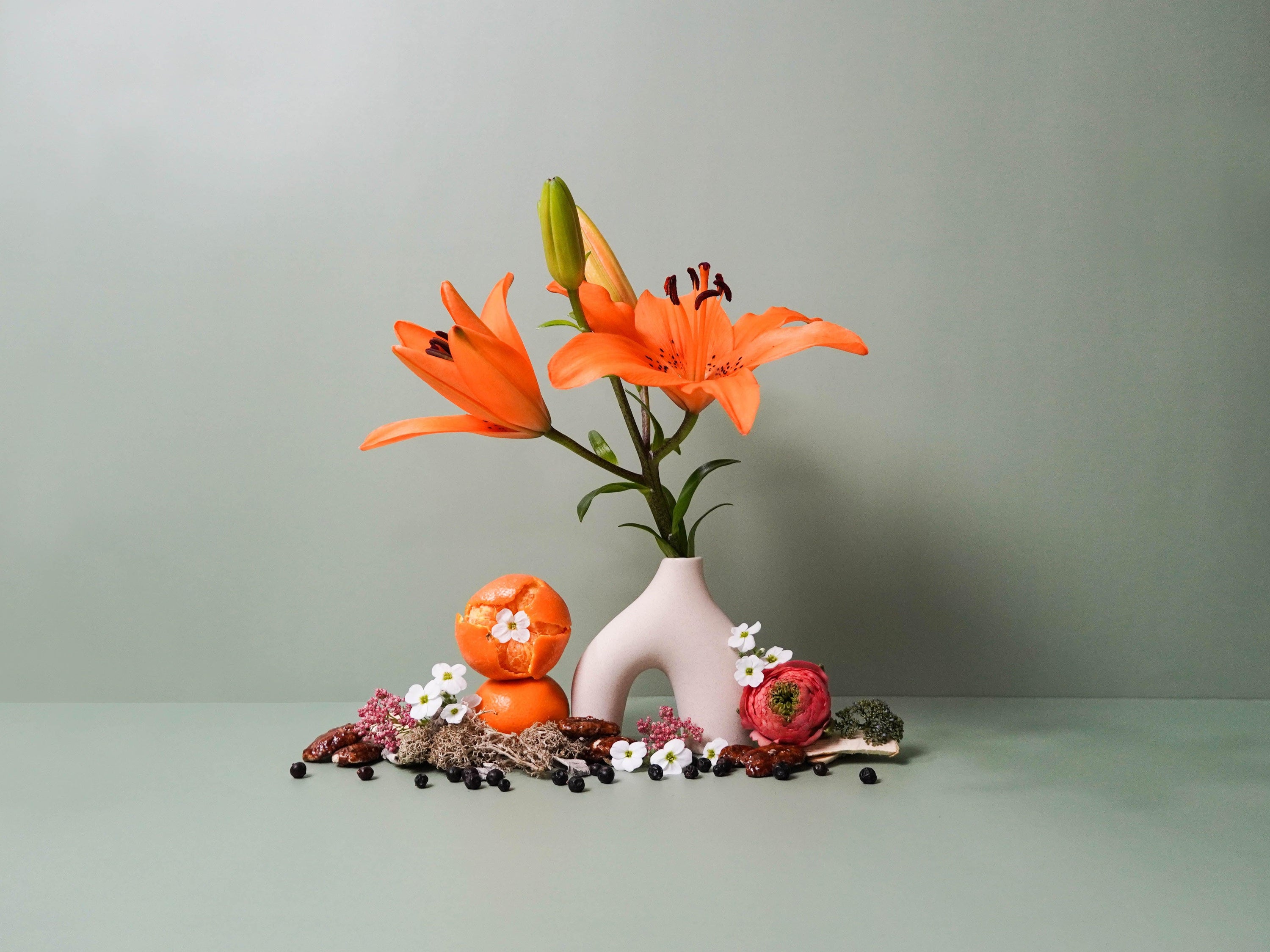 A minimalist still life composition featuring vibrant orange lilies in a modern white ceramic vase, surrounded by scattered botanicals and textures. The arrangement includes a halved orange, delicate white and pink flowers, green moss, dark juniper berries, and earthy spices. Set against a muted sage green background, the scene evokes a balance of bold colour and serene natural elements.