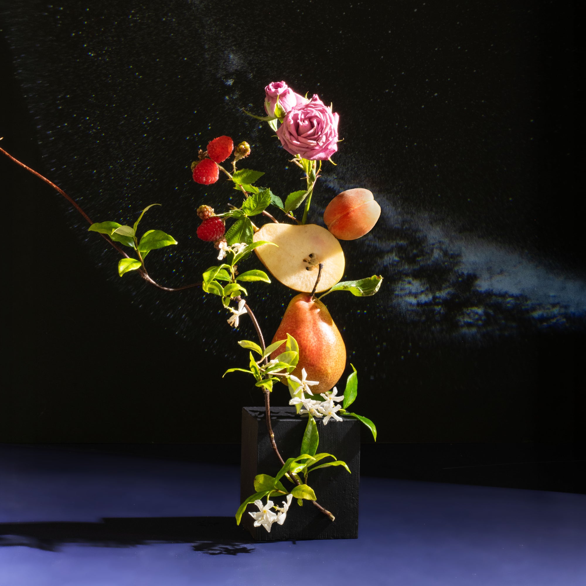 A vertical fruit and flower arrangement in a black square vase against a dark studio background. The composition includes a pink rose, red raspberries, a sliced pear, a whole pear, a peach, green leaves, and small white flowers.