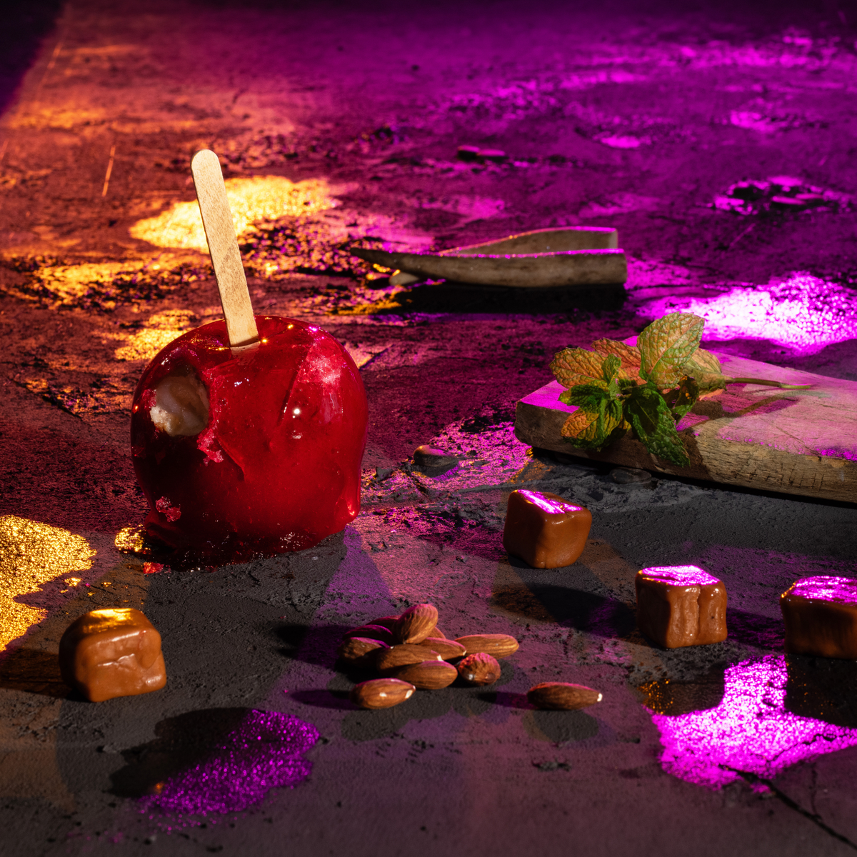 Red candy apple surrounded by caramel candies, almonds and fresh mint on a dark textured surface with dramatic pink and golden lighting.