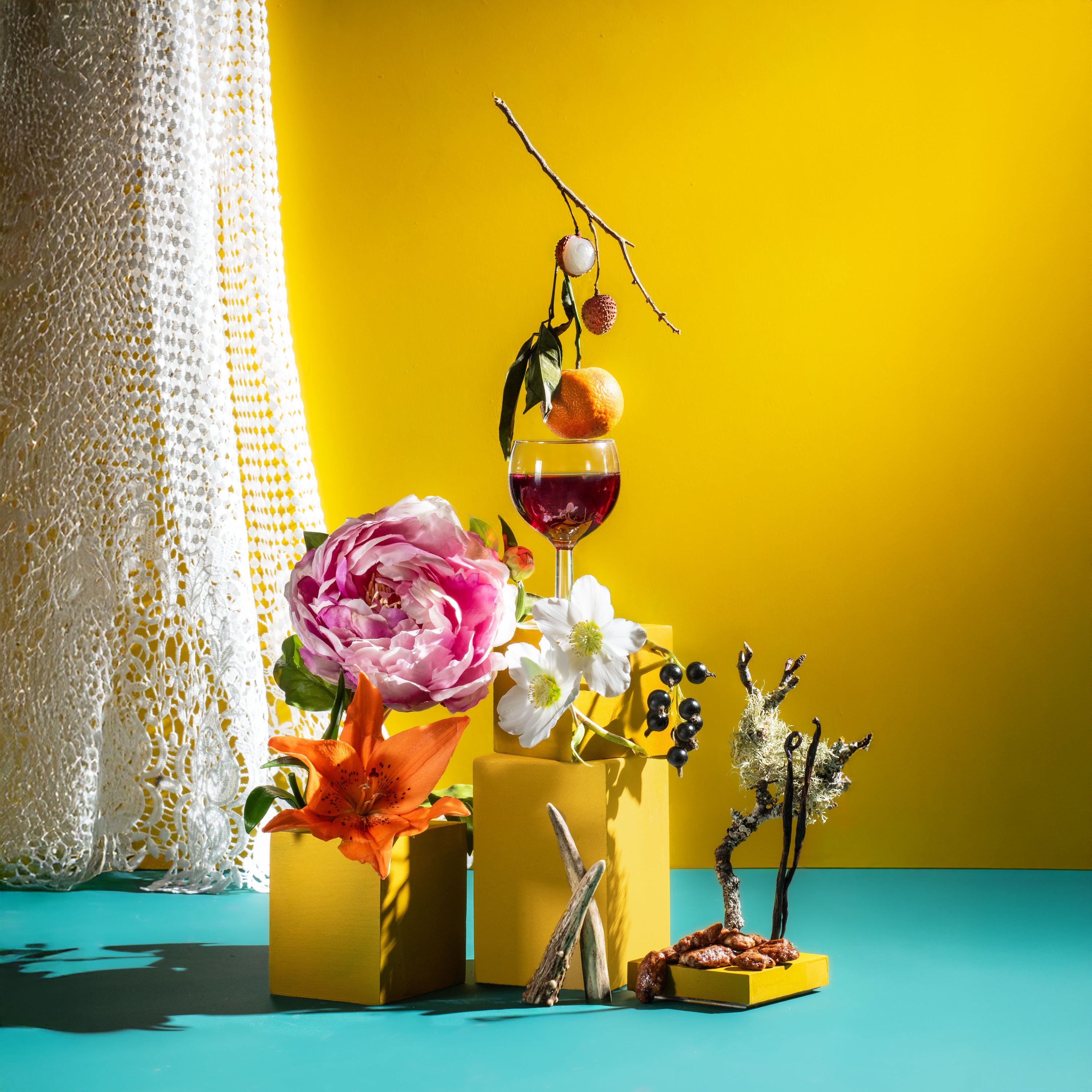 Decorative setup with flowers, a glass of red wine, and branches against a yellow wall.