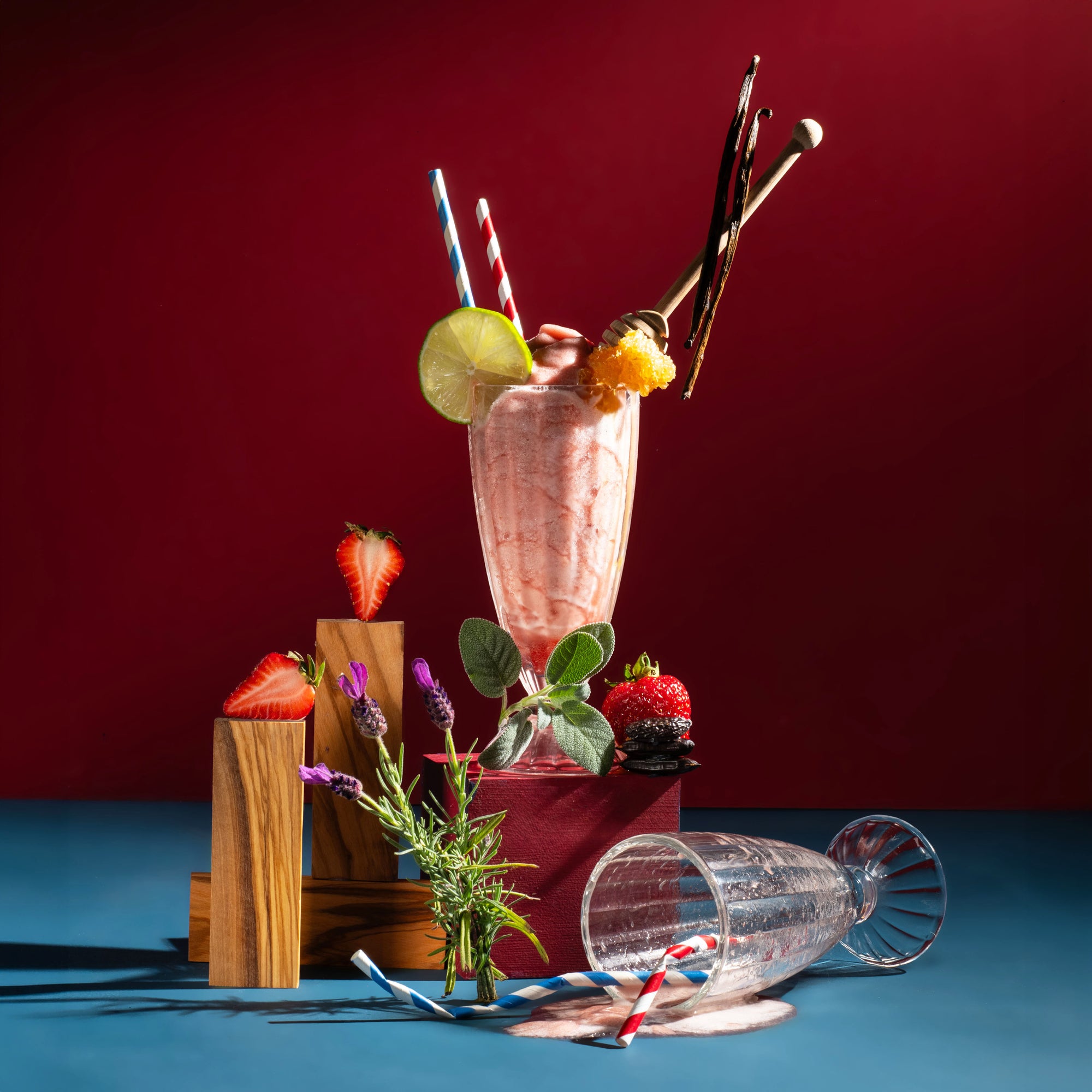A pink milkshake in a tall clear glass with a lime wheel, orange slice, and striped straws, styled on a blue surface against a dark red background with strawberries, flowers, a tipped-over glass, and wooden blocks.