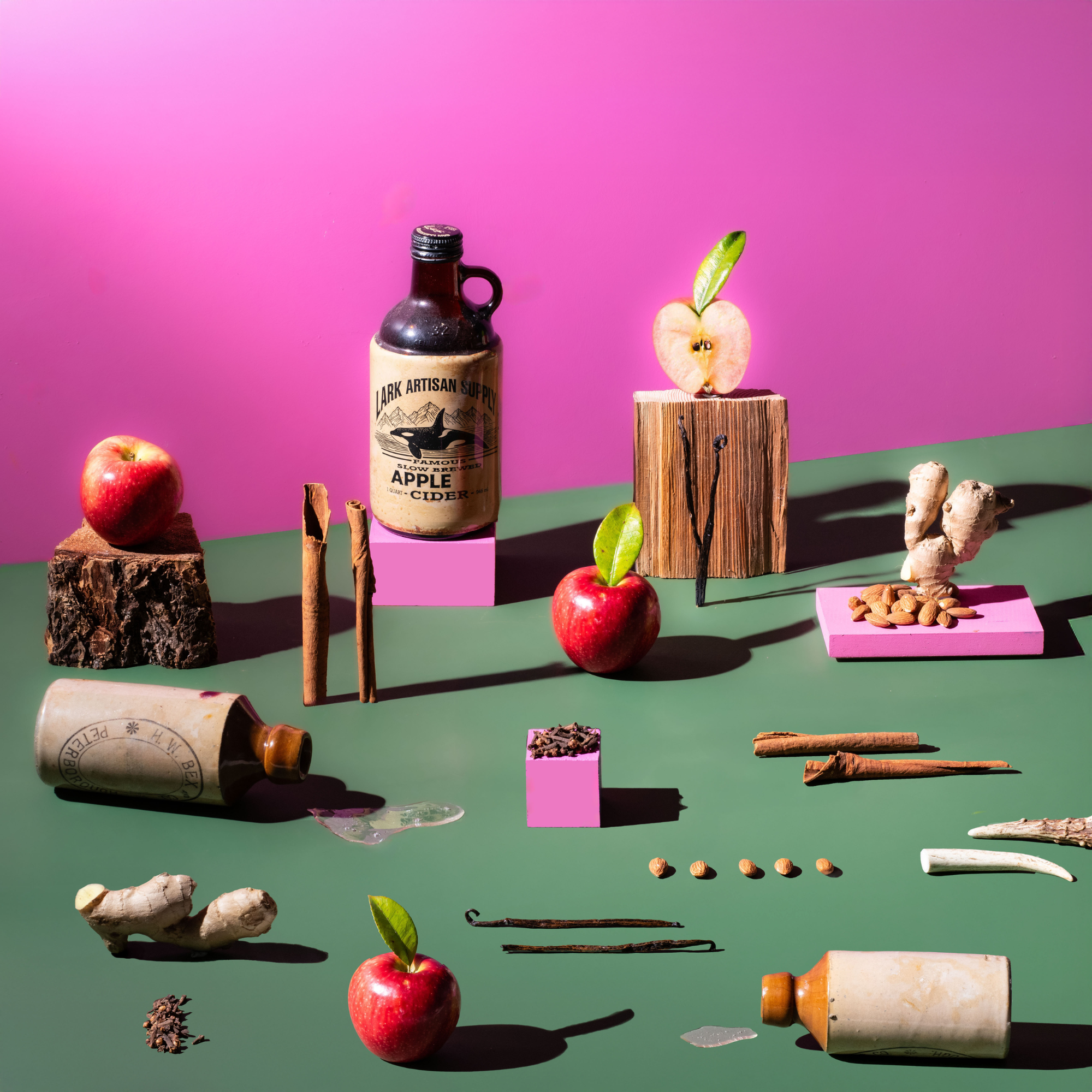 Still life on a green surface with a pink backdrop, featuring apples, cinnamon sticks, vanilla pods, ginger, almonds, and Lark “Apple Cider” bottles arranged on wood blocks.