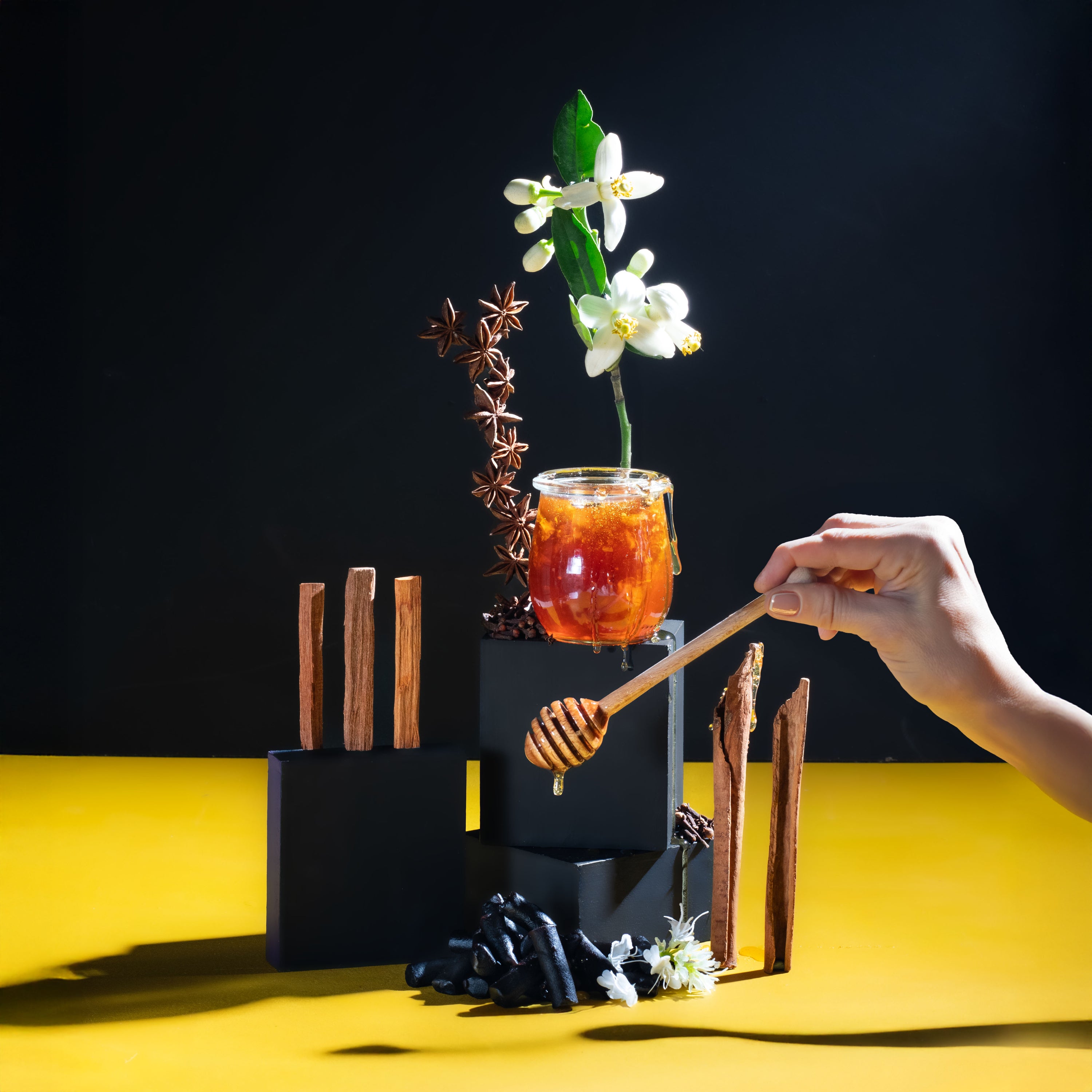 Hand holding a honey dipper over a glass of amber liquid with flowers and spices on a yellow and black background