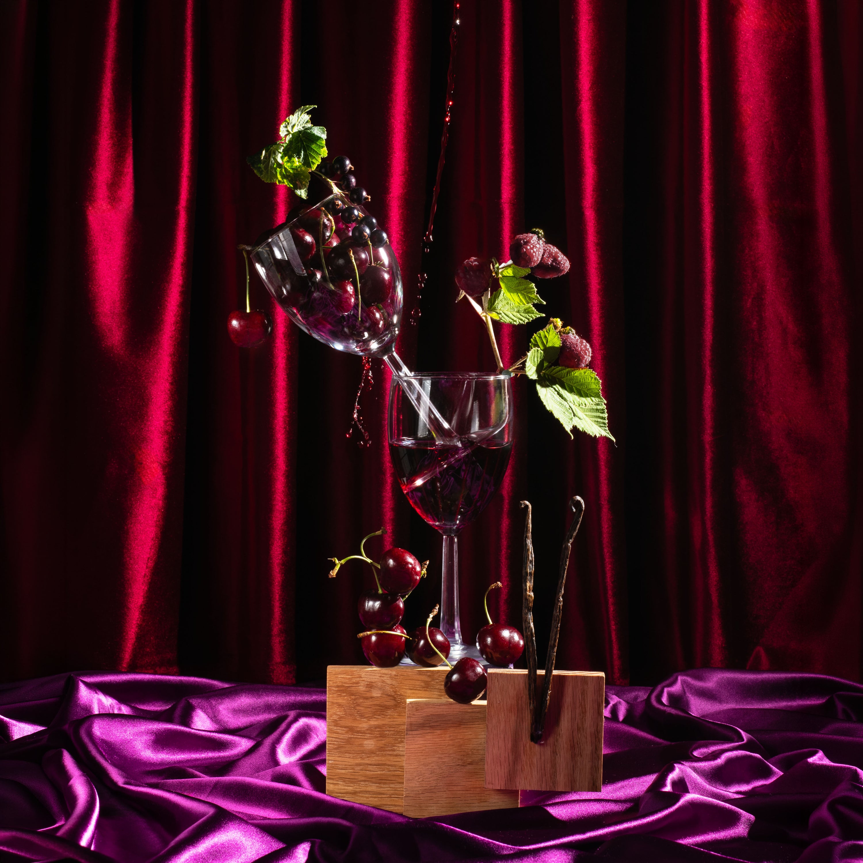 A still life arrangement of red wine in stemmed glasses, cherries, green leaves, and dark chocolate on a wooden block, set against a deep red velvet curtain and magenta satin fabric.