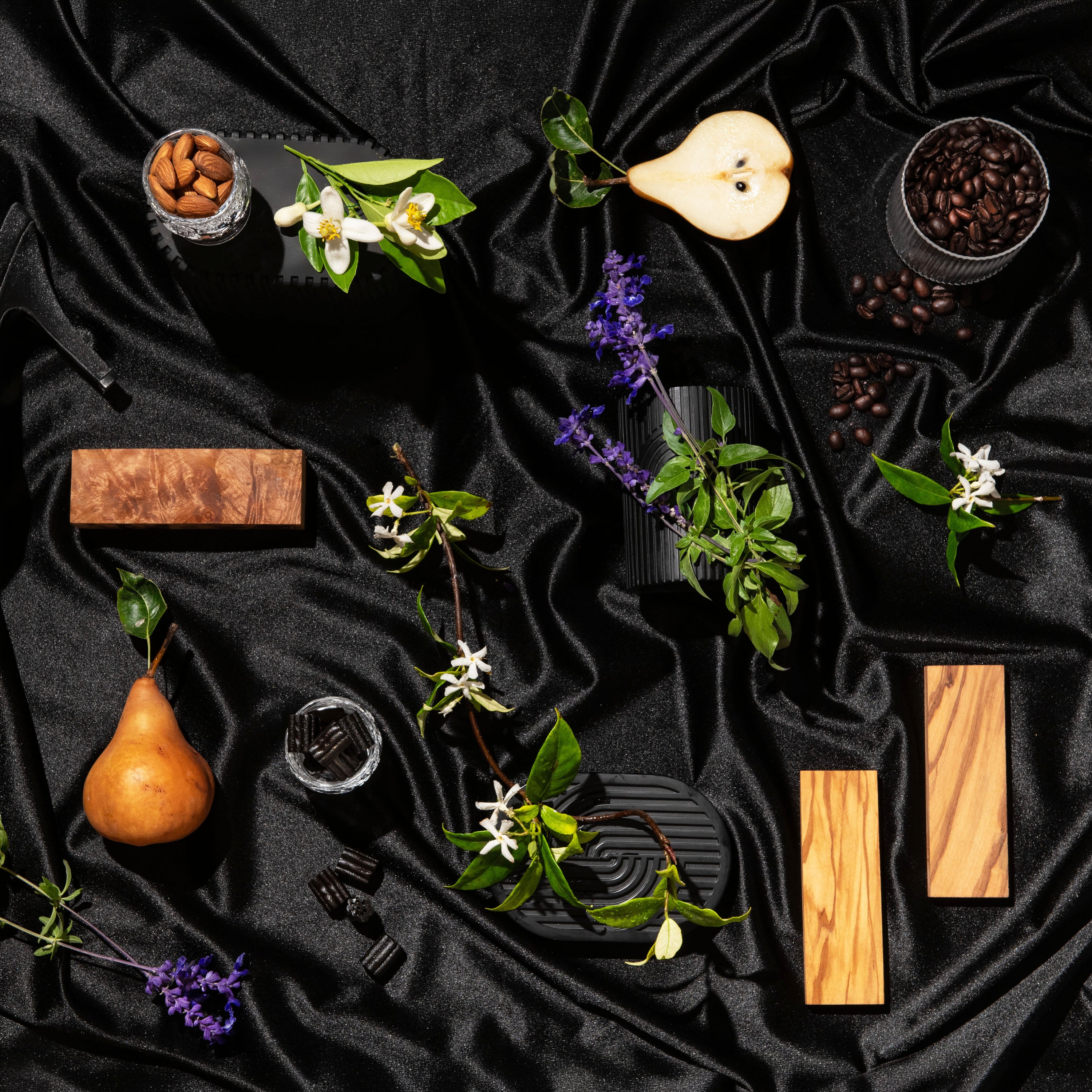A styled flat-lay of several rectangular wooden soap bars with different wood grain patterns, arranged on a draped black satin fabric with decorative props including a pear, coffee beans, flowers, and small glass containers.