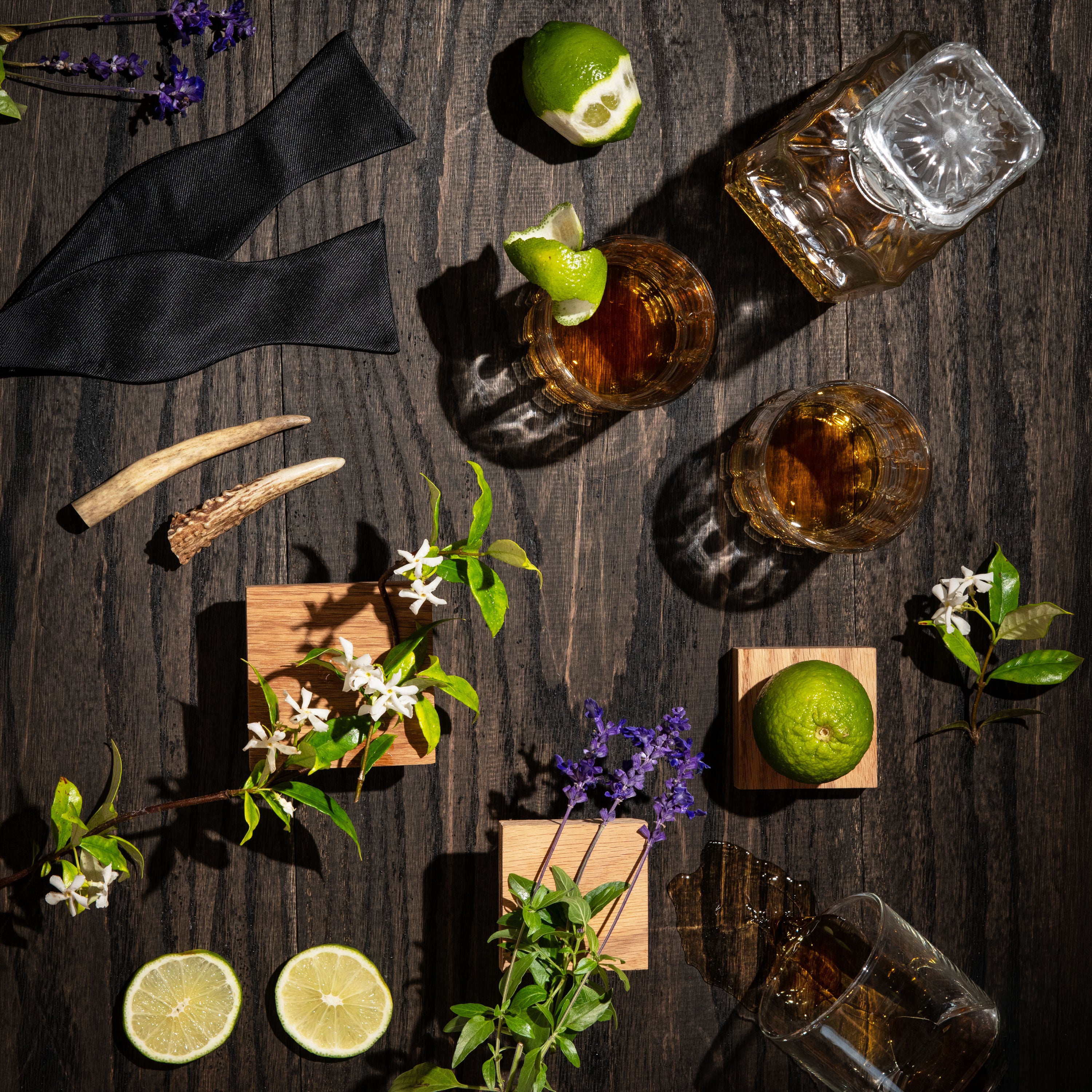 An overhead styled flat-lay on a dark wood surface featuring two rocks glasses filled with amber liquor, a cut lime garnish, a clear cut-glass tumbler, a black cloth, small horns, flowers, and lime slices arranged decoratively.