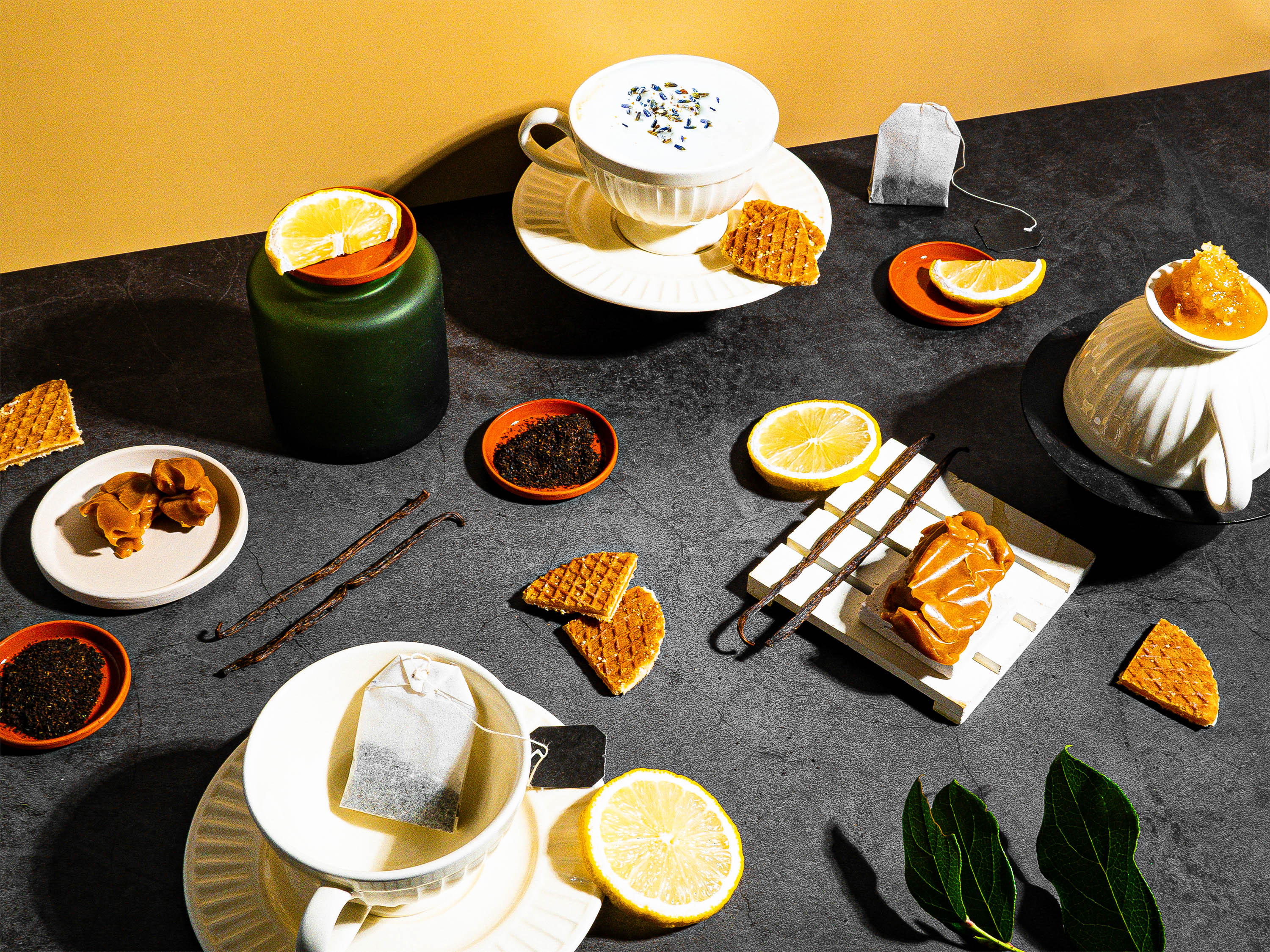 A vibrant, tea-inspired flat lay set against a dark, textured backdrop with a warm golden accent. The scene includes delicate tea cups, one topped with floral garnish and another with marmalade, alongside stroopwafels, lemon slices, caramel swirls, loose tea leaves, and vanilla beans. A green jar adds depth, while sprigs of fresh green leaves bring a natural element to the composition. Perfectly balanced, the setup evokes cozy indulgence with a sophisticated touch of citrus and sweetness.