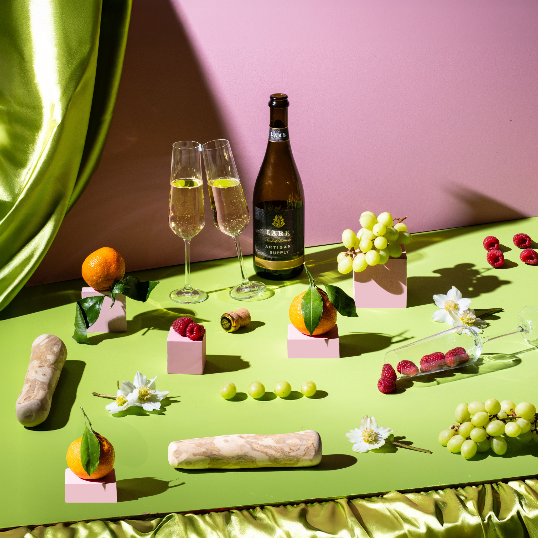 Bottle of champagne, two glasses, and assorted fruits and pastries on a green surface with a pink background.