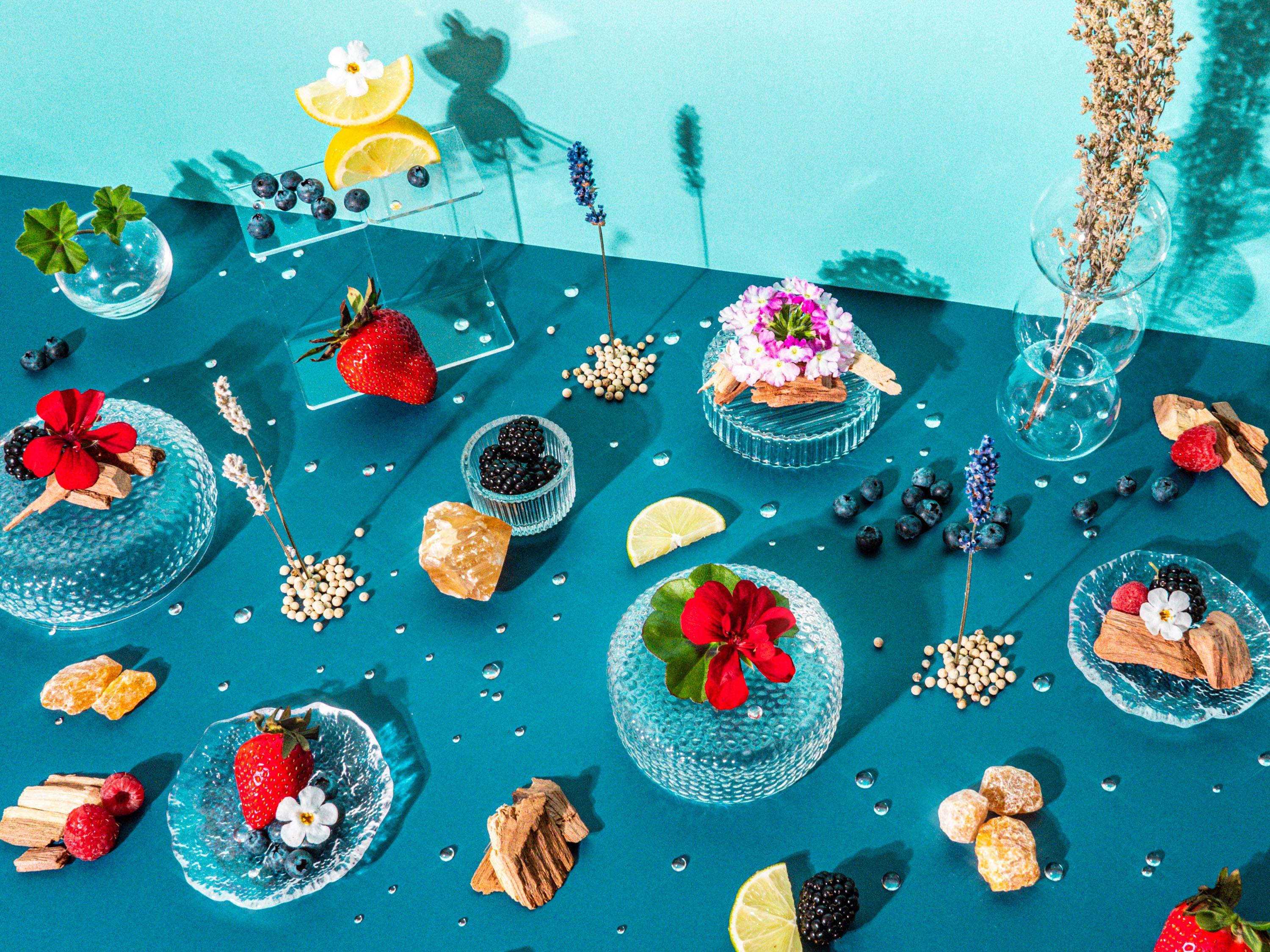 A delightful and artistic display of botanical and culinary elements on a teal background. The scene includes various fresh berries like blueberries and strawberries, slices of lemon, and vibrant flowers, all artistically arranged on textured glass plates and alongside pieces of wood and dried herbs. The composition is accentuated by glass vases holding sprigs of lavender and other dried botanicals, creating a fresh, vibrant tableau that evokes a sense of summer freshness and natural beauty.