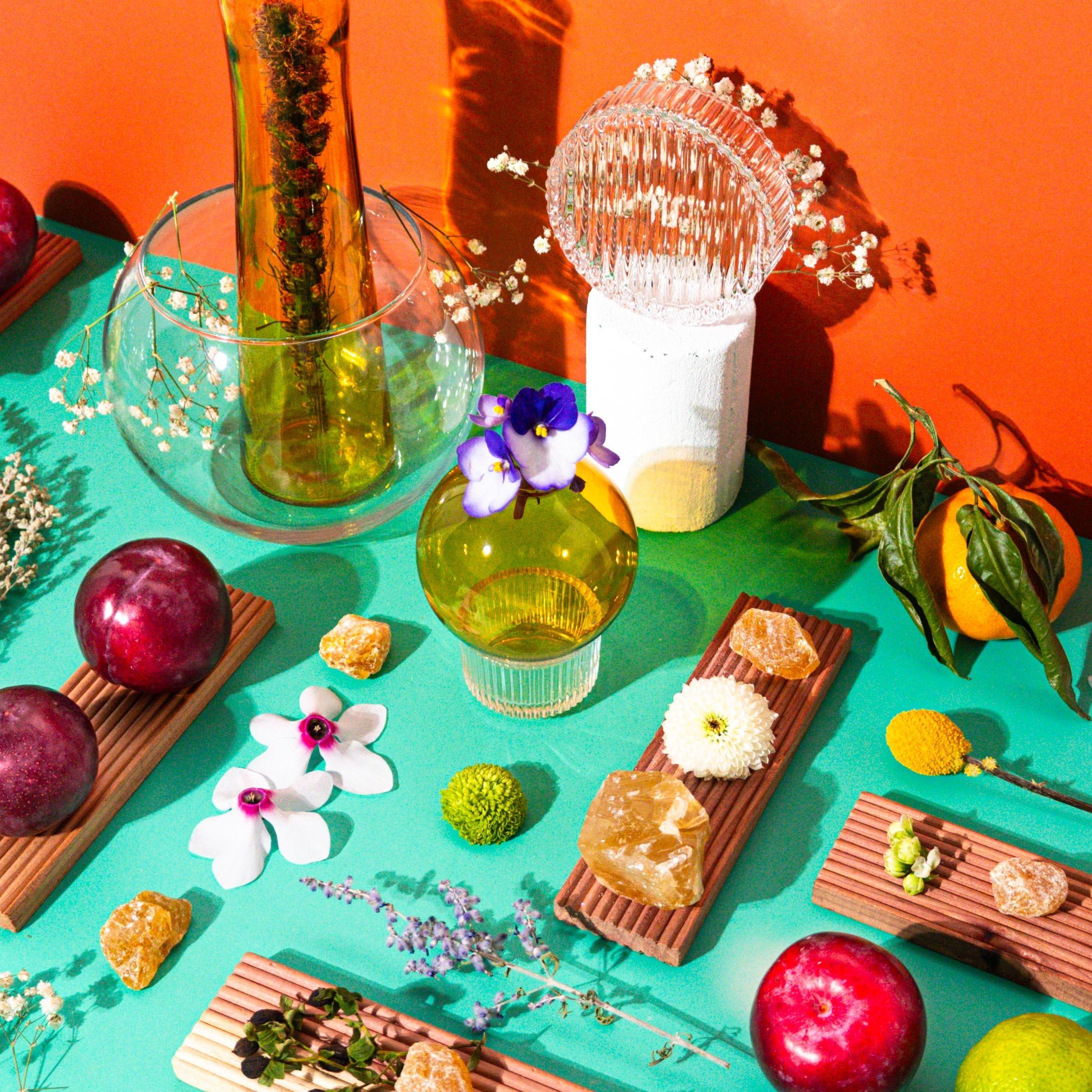 An arrangement showcasing a bottle of fragrance oil with citrus fruits, plums, and flowers displayed around it on a colorful background.