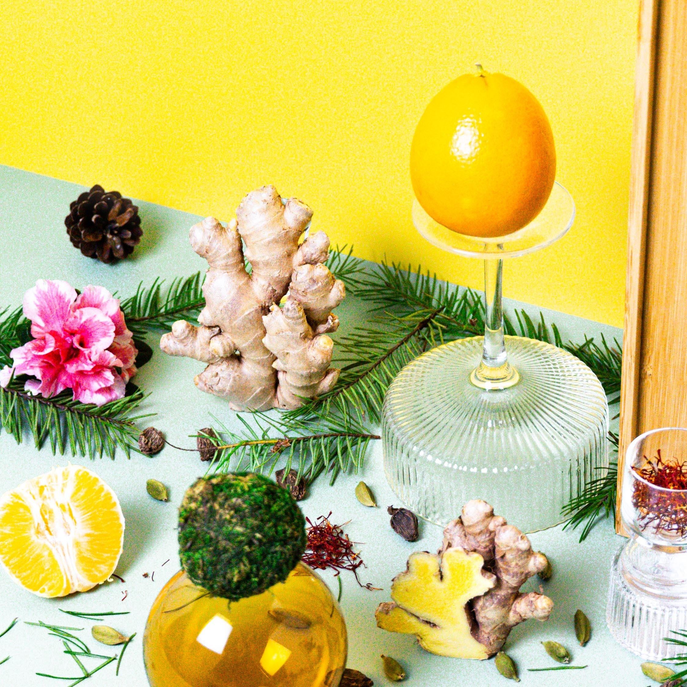 A still life arrangement featuring a bottle of fragrance oil, with decorative elements such as citrus fruits, herbs, and flowers, against a yellow background.