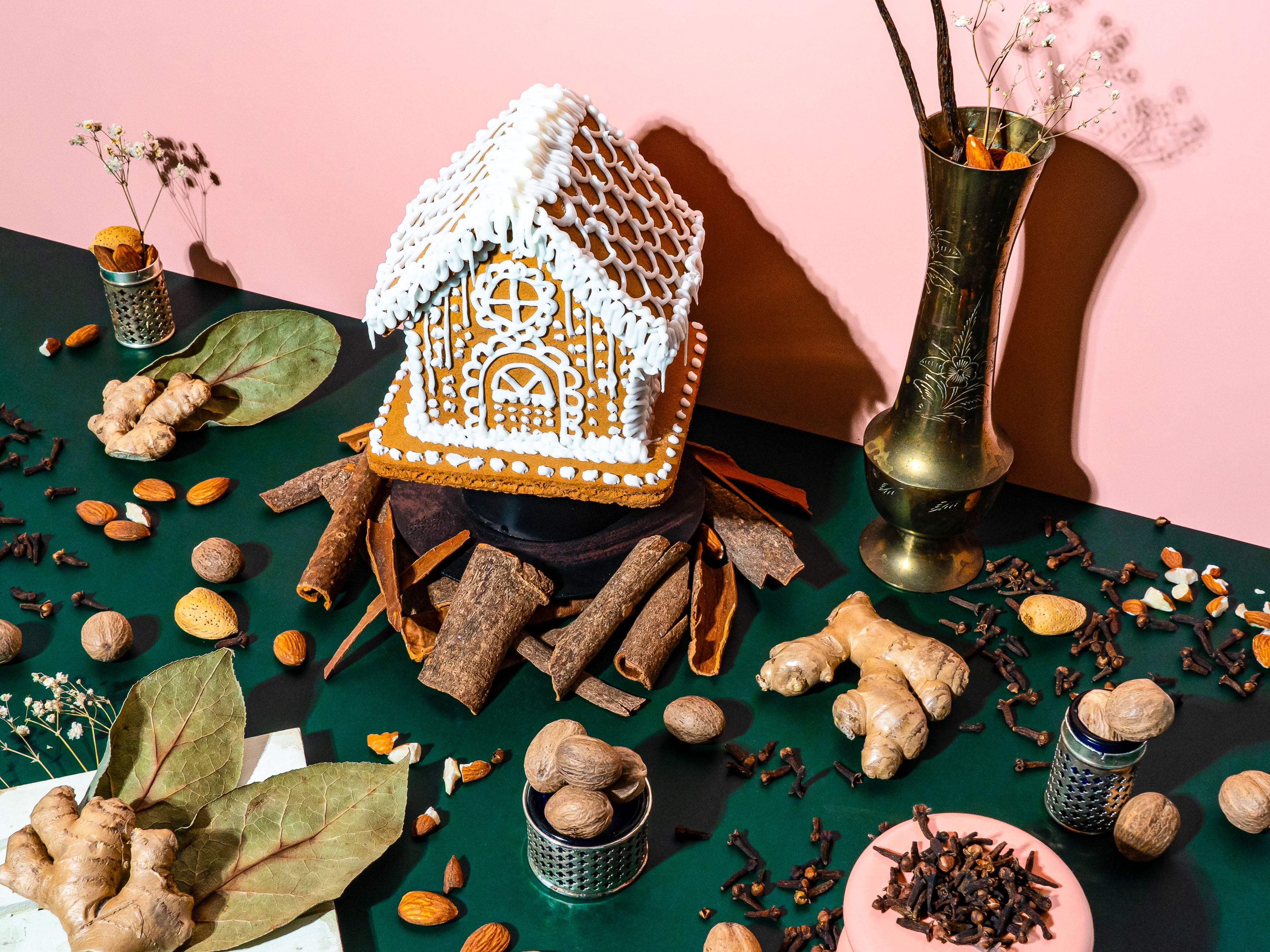 A gingerbread house with intricate white icing details sits atop a dark pedestal, surrounded by an array of warm, aromatic spices and nuts. Cinnamon sticks, whole cloves, nutmeg, and ginger roots are scattered across a deep green surface, evoking rich, spiced scents. Almonds and walnuts add texture, while a vintage brass vase holds vanilla pods and dried botanicals. The pink backdrop contrasts with the earthy tones, creating a striking, cozy scene reminiscent of festive baking and warm winter gatherings.