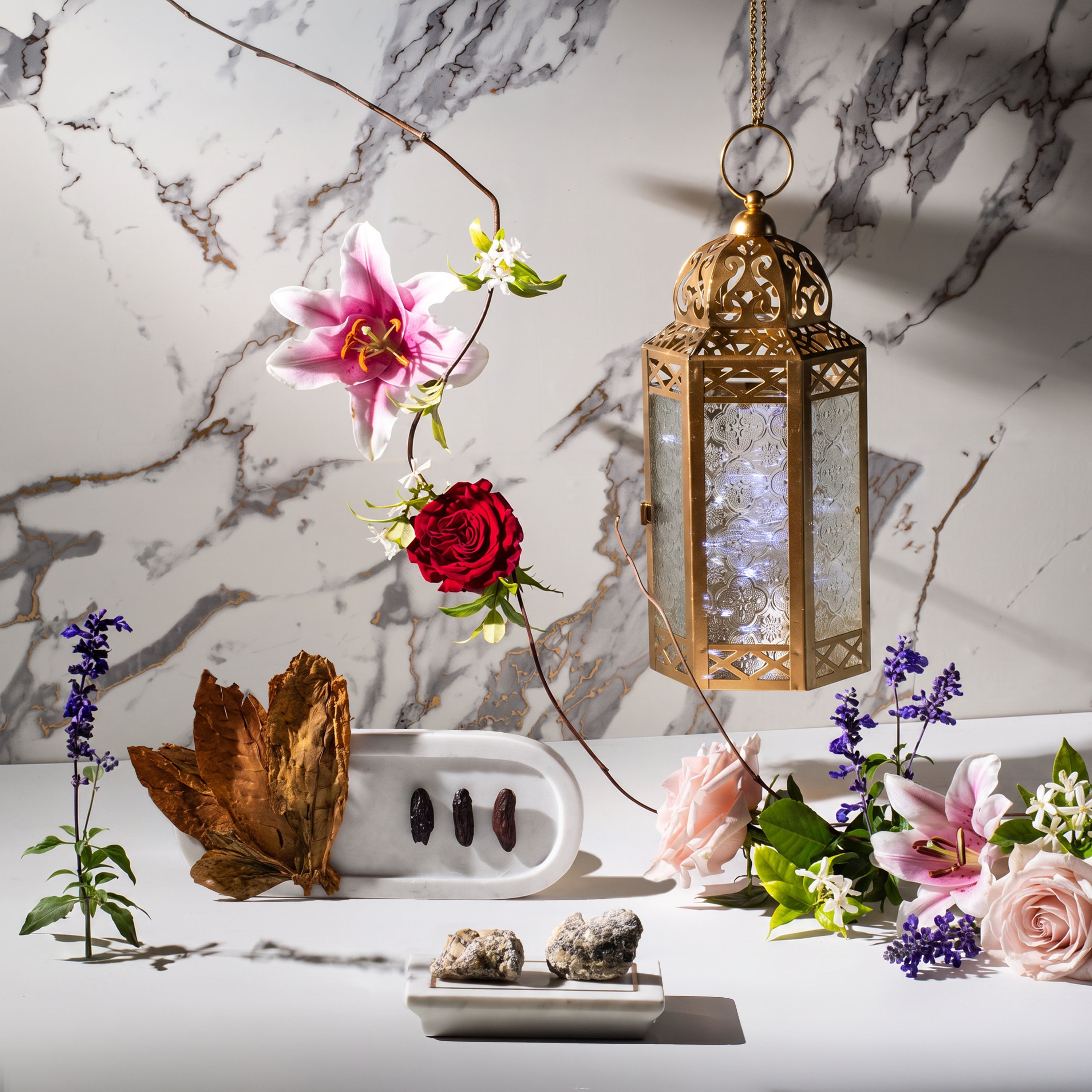 Elegant still life with pink lilies, red and blush roses, lavender, jasmine blossoms, oud wood, dates, decorative lantern, and marble backdrop.