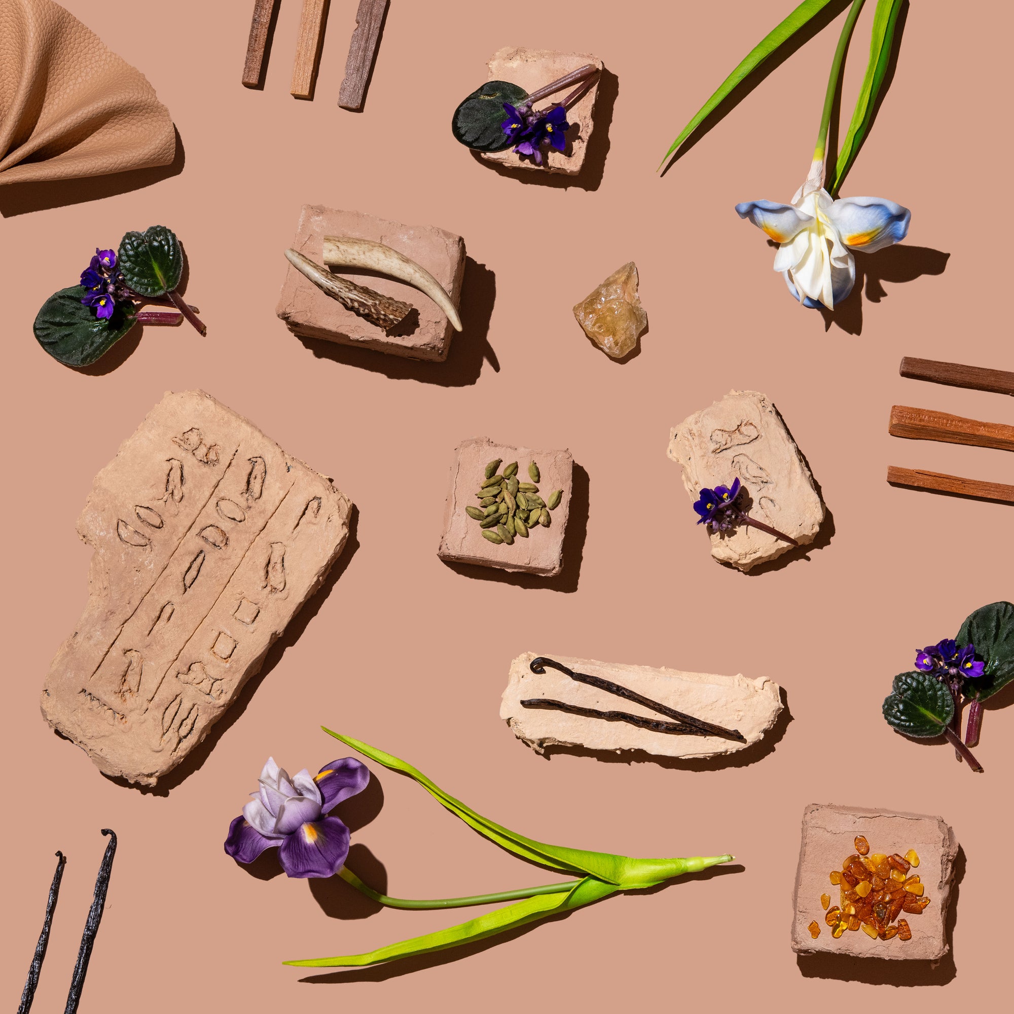 Flat lay on a warm tan background with clay tablets stamped with hieroglyphs, surrounded by vanilla beans, cardamom, amber resin, wood strips, and purple flowers.