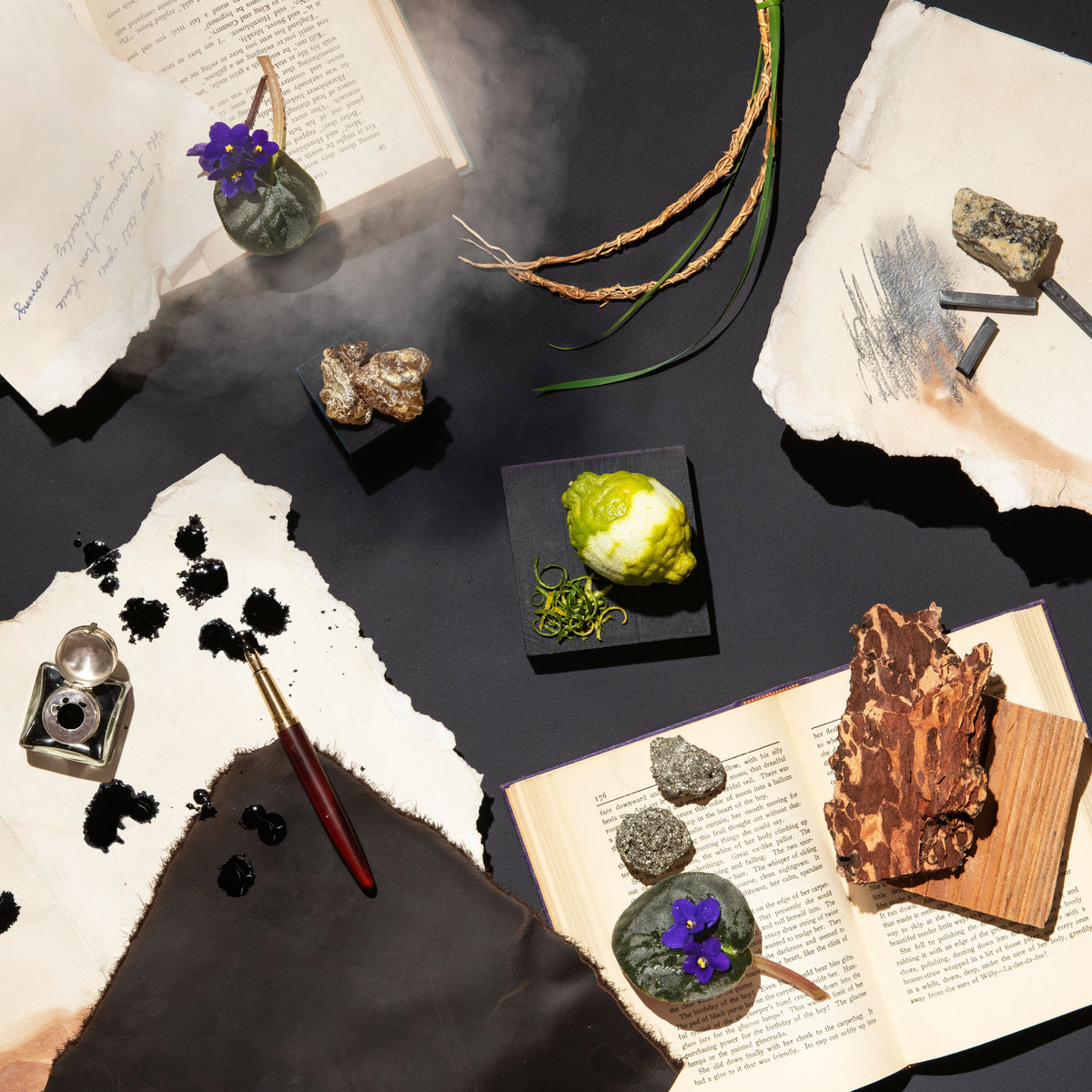 An overhead styled flat-lay featuring a small green glass vase with a purple flower, natural rock specimens, torn white paper, ink, a paintbrush, and dried botanical elements arranged on a dark surface.