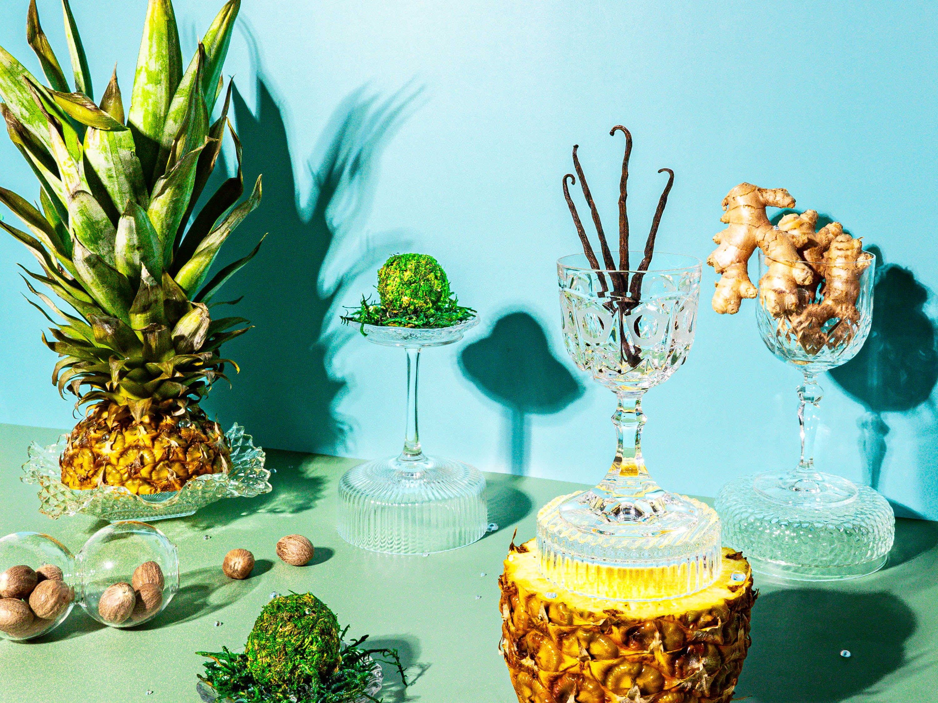 A vibrant tropical display with a fresh pineapple, vanilla beans, ginger roots, moss spheres, and scattered nutmeg seeds, arranged in elegant crystal glassware against a turquoise background.