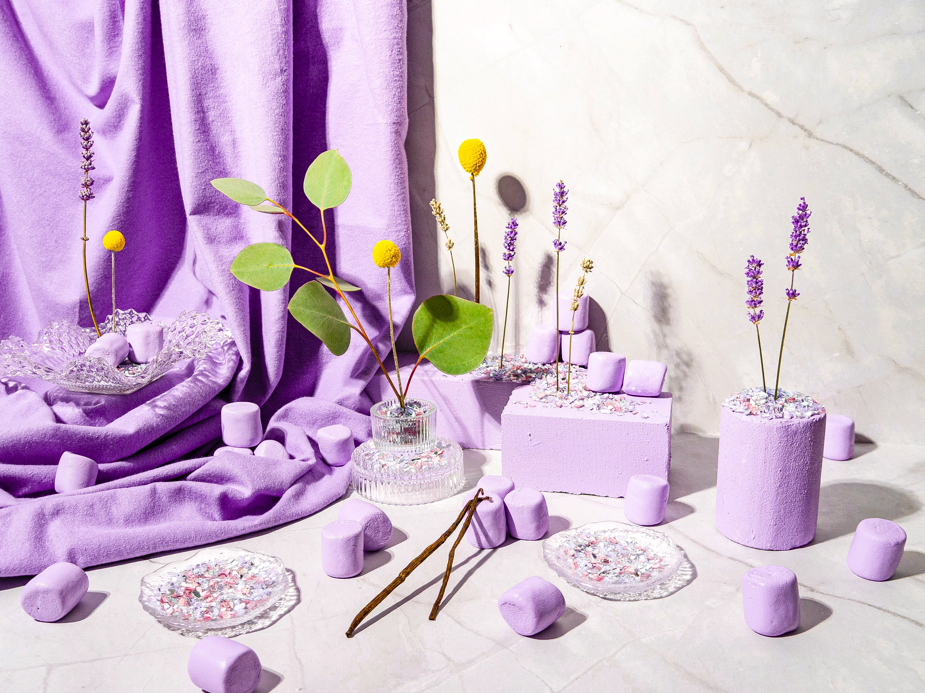 A whimsical and delicate display featuring soft lavender hues. Lavender stalks, golden billy buttons, and fresh green leaves are set in glass vases and lavender-colored blocks sprinkled with confetti-like textures. Plush lavender marshmallows and draped fabrics create a cozy, dreamy atmosphere, complemented by crystal plates and scattered decorative elements on a marble surface. The scene exudes a balance of softness, elegance, and playful charm.