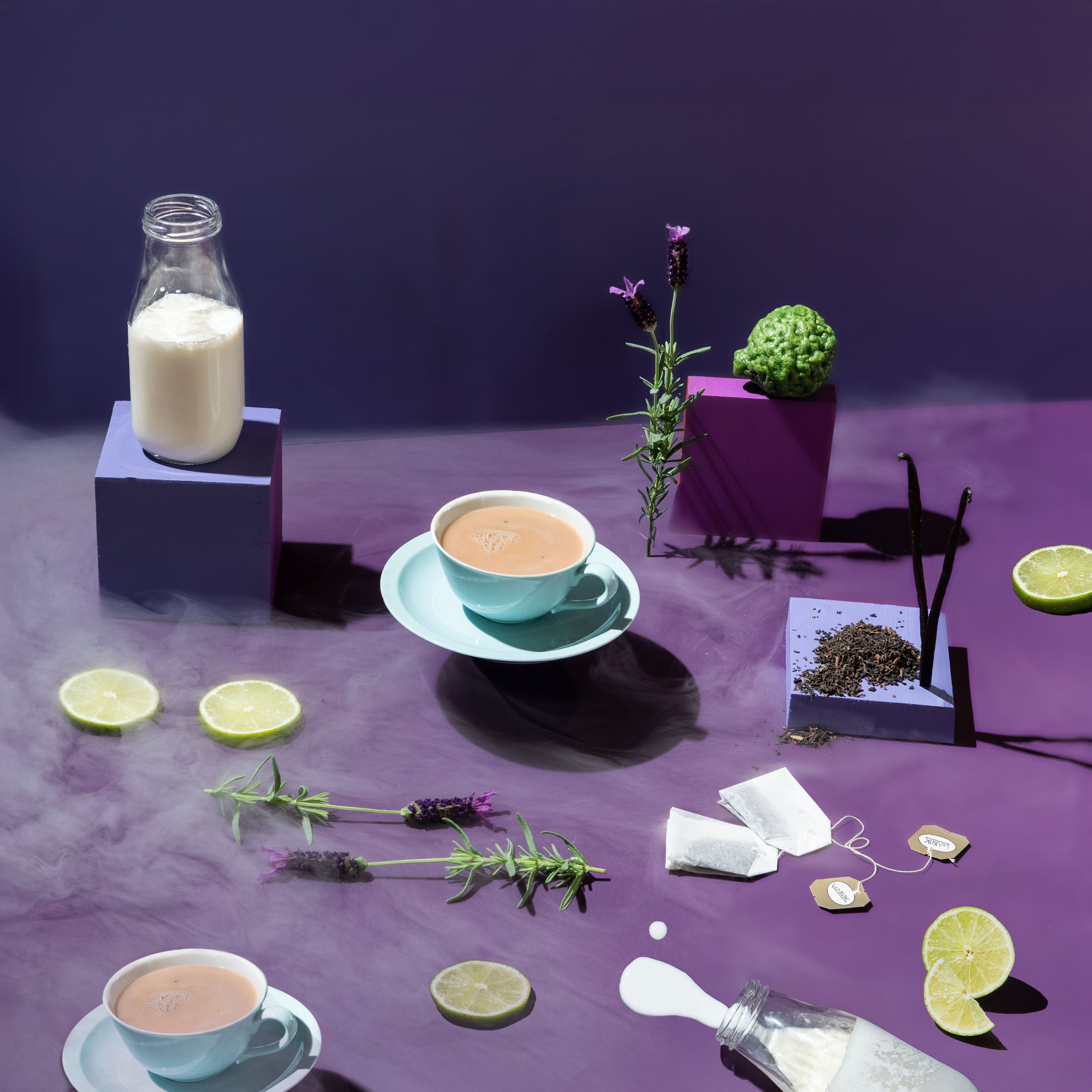 Tea setup with cups, milk bottle, lime slices, and lavender on a purple surface.