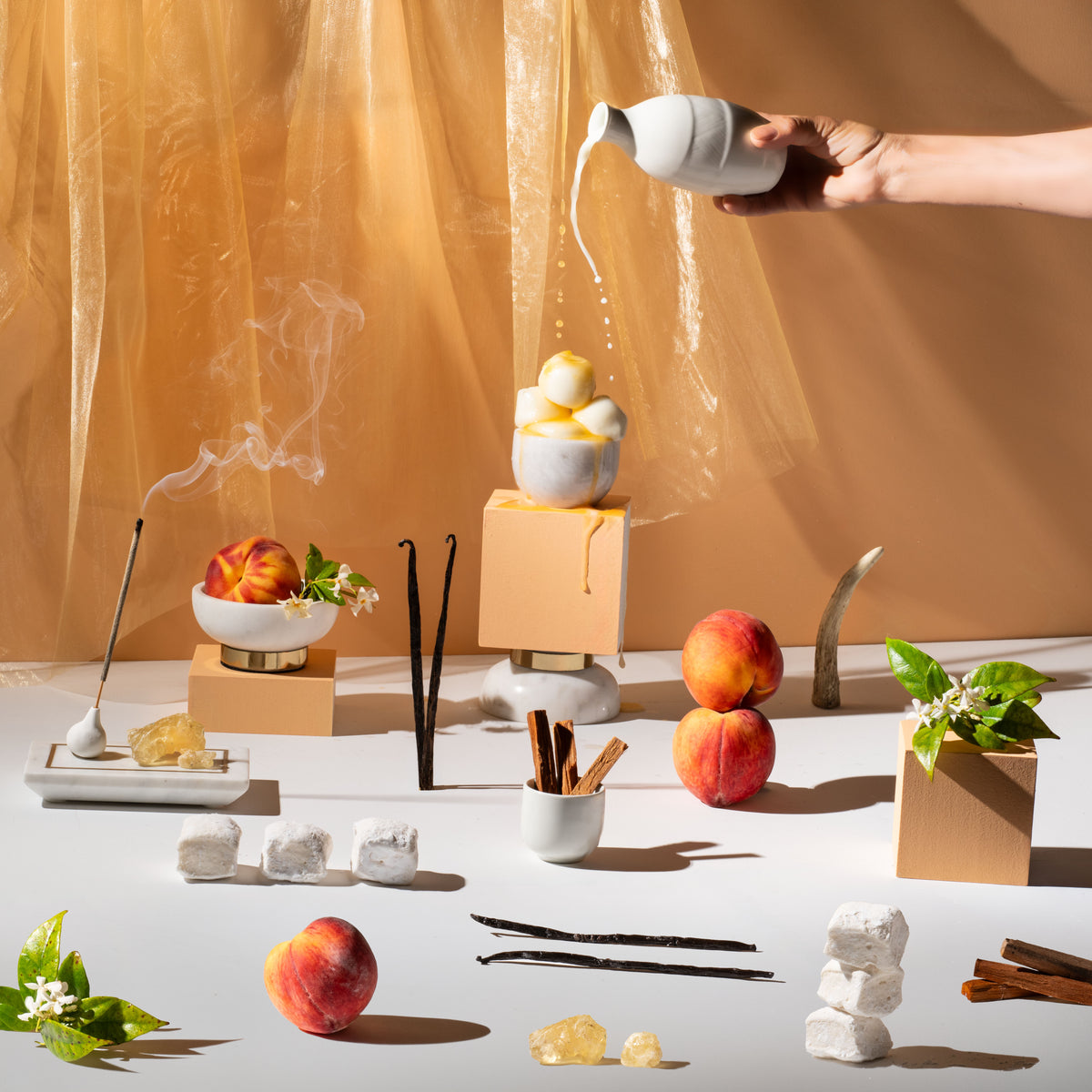 A styled still life of ceramic objects and food props on a white surface against a warm tan draped backdrop. A hand is pouring liquid from a white ceramic bottle into a small white dish containing yellow fruit pieces.