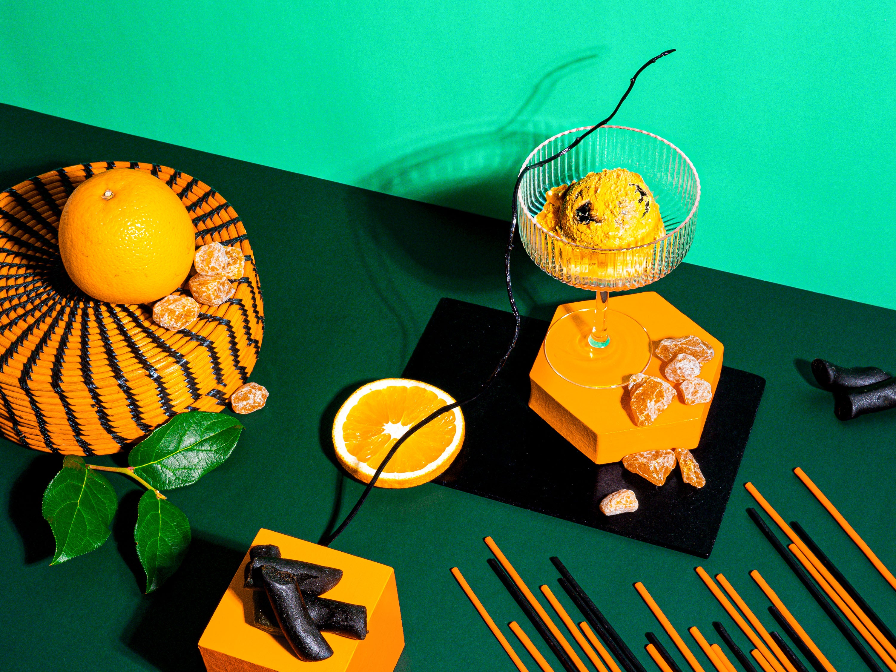 A striking arrangement featuring vibrant oranges, black licorice pieces, and crystallized orange candies. A scoop of orange and licorice-inspired dessert sits elegantly in a glass atop geometric orange blocks. Set against a rich green backdrop, the scene is accented with a woven basket, fresh green leaves, and sleek black incense sticks, evoking bold contrasts and indulgent sophistication.