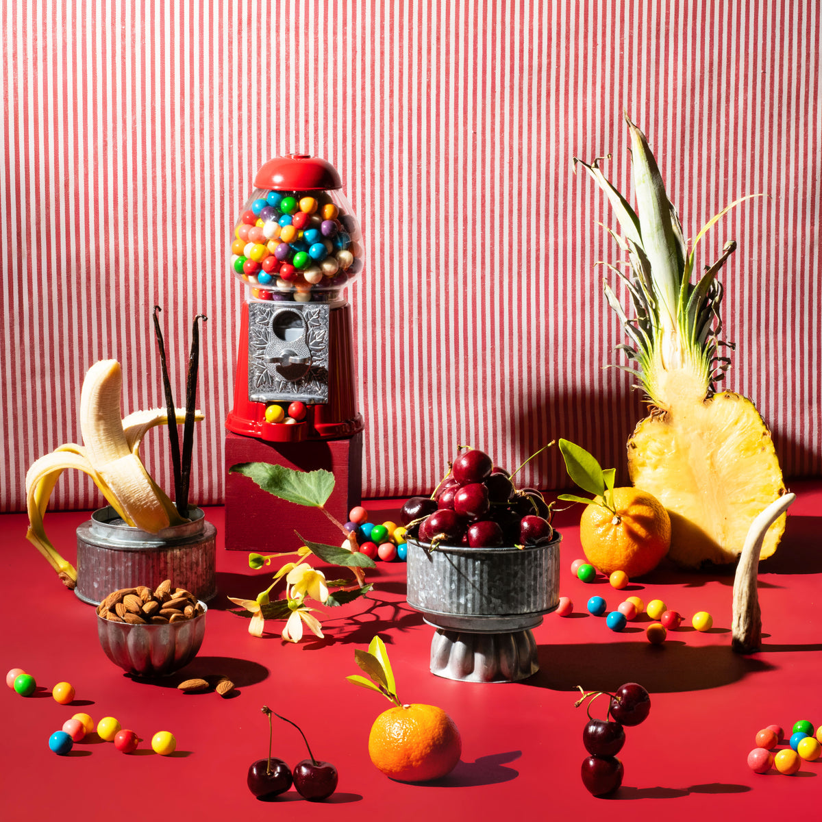 A styled still life scene with fruit and candy props against a red-and-white striped backdrop: a pineapple, banana, cherries, an orange, nuts, and a red gumball machine filled with colorful candies on a red surface.