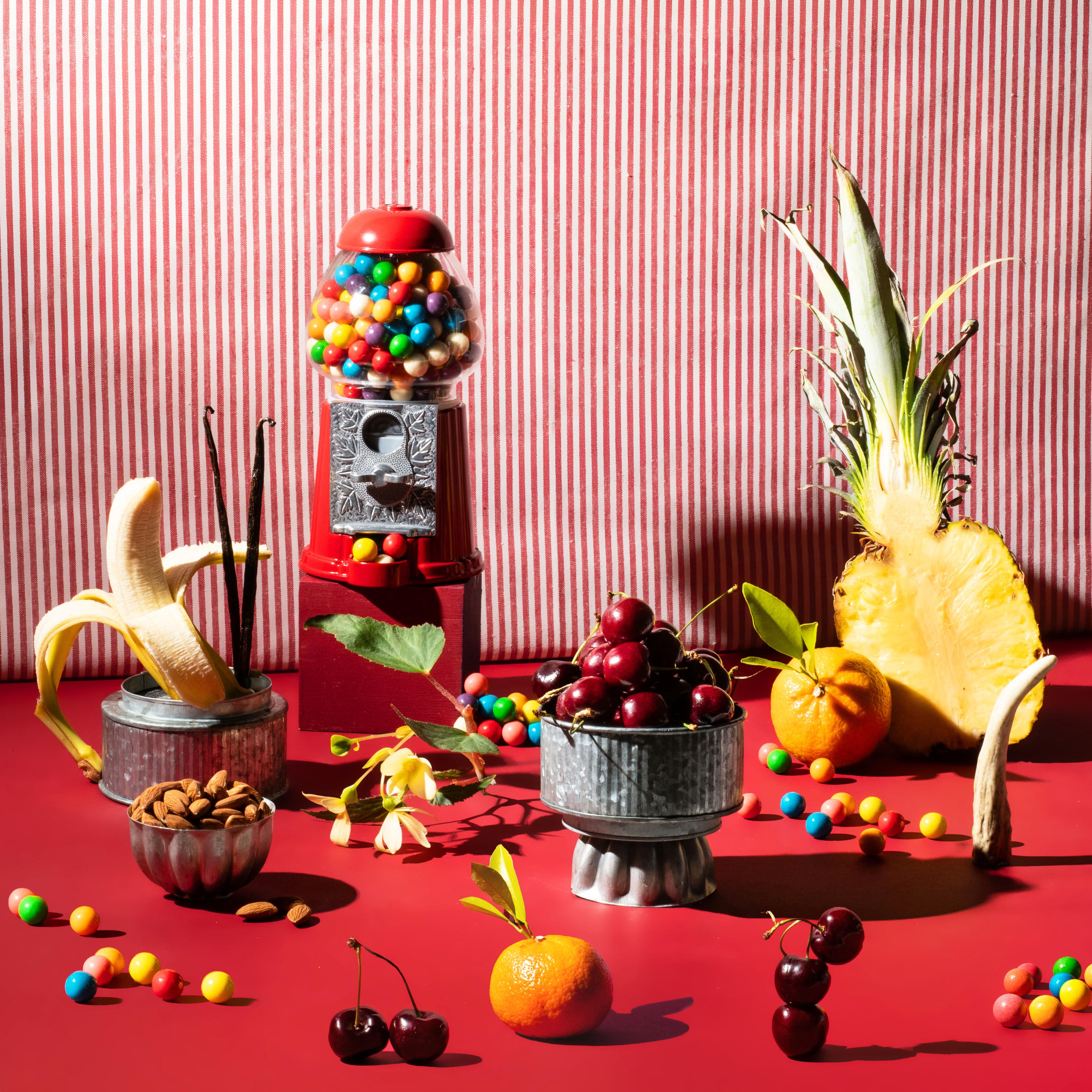 A styled still life scene with fruit and candy props against a red-and-white striped backdrop: a pineapple, banana, cherries, an orange, nuts, and a red gumball machine filled with colorful candies on a red surface.