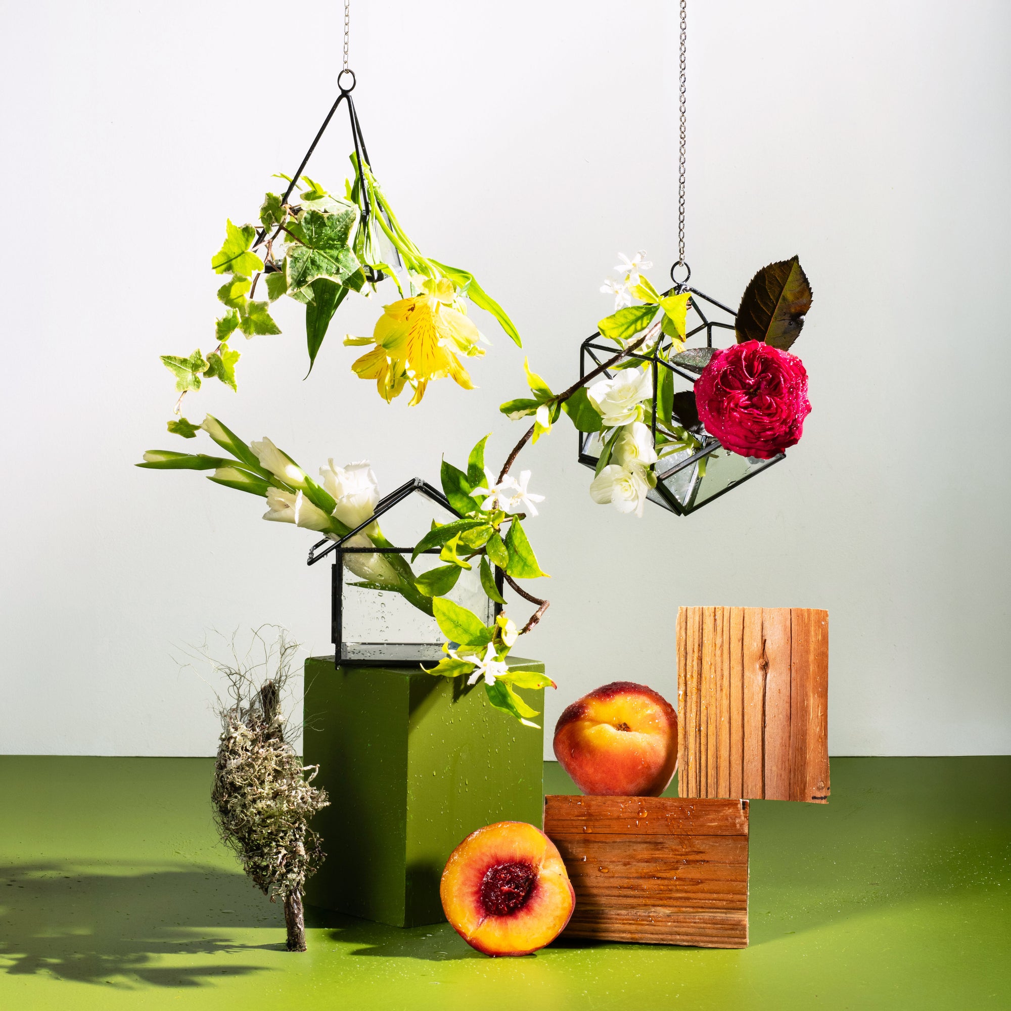 Fresh floral still life with yellow alstroemeria, white roses, ivy, jasmine vine, peaches, cedarwood blocks, moss, and hanging glass terrariums on a green surface.