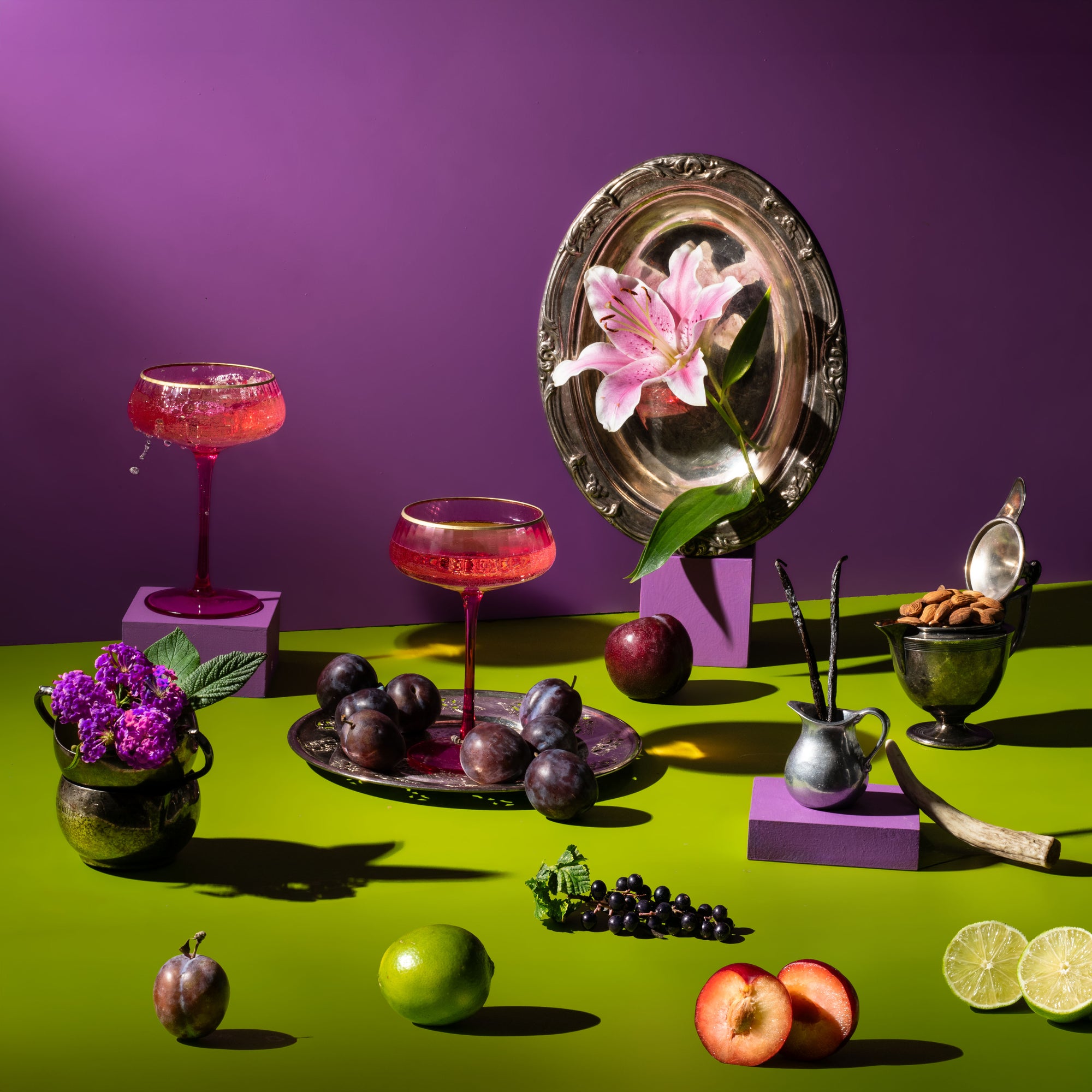 A stylized still life arrangement on a bright green tabletop against a purple backdrop, featuring pink cocktails in coupe glasses, plums, limes, sliced apples, purple flowers, a silver plate with a pink lily, and small decorative vessels.