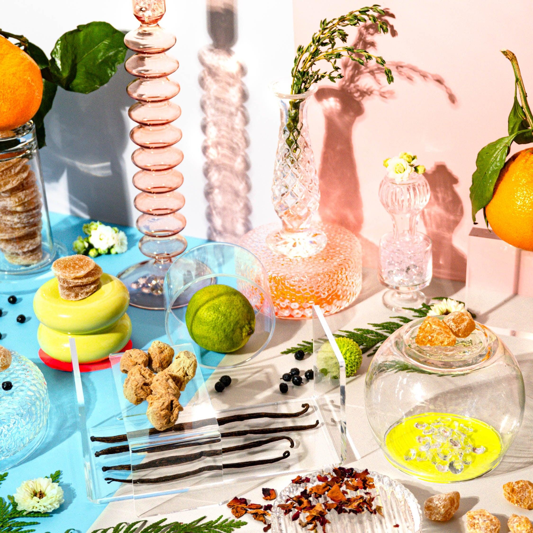 A still life arrangement featuring various elements associated with the product title, such as a rose, ginger crystals, cedarwood, and vanilla, along with other decorative items like citrus fruits and glass bottles.