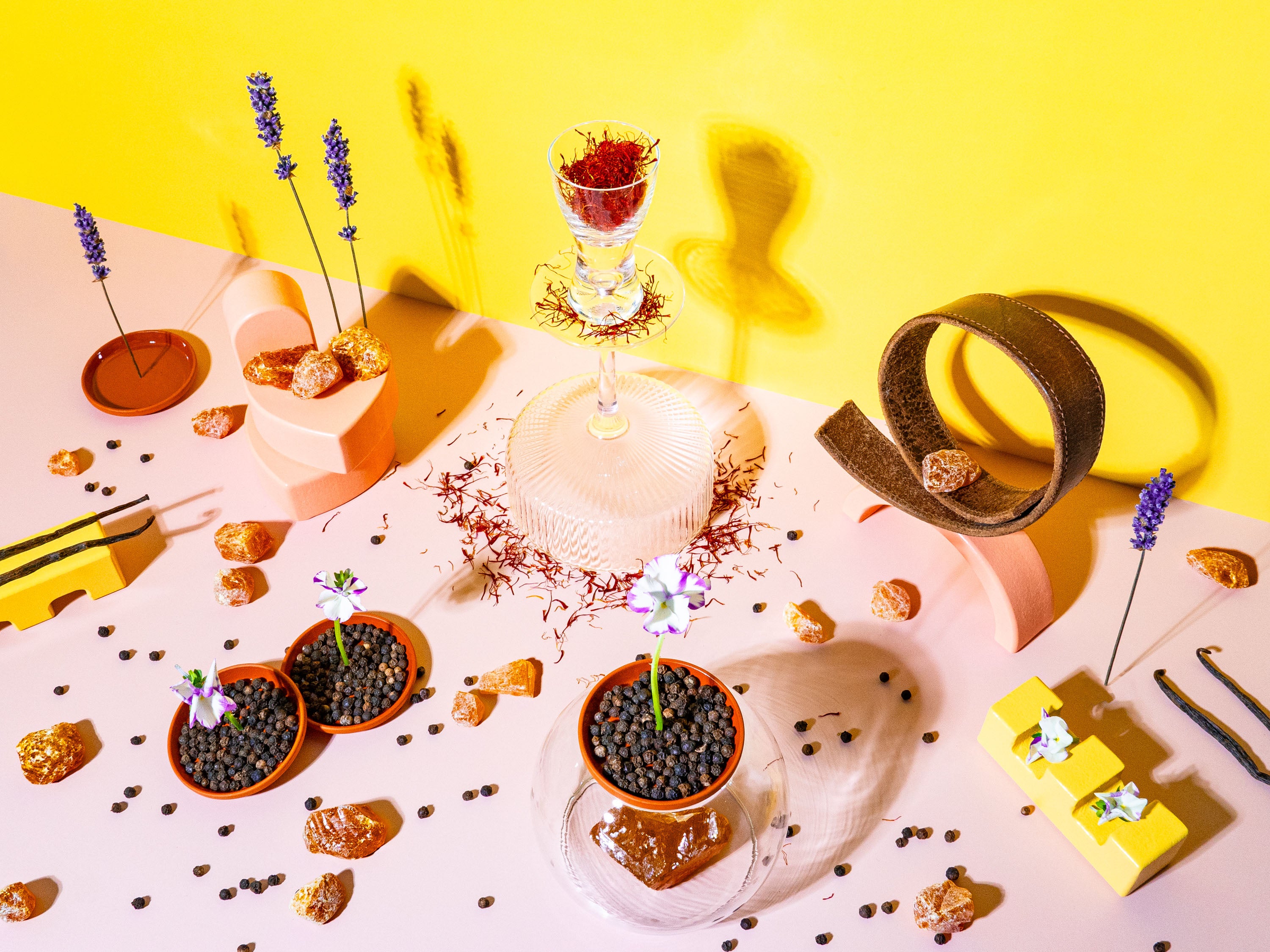 A vibrant and artistic tabletop display on a yellow background featuring a variety of objects: small terracotta pots with black peppercorns, delicate purple flowers, geometrically shaped wooden sculptures, and amber-colored rocks scattered across a pink surface. In the center, a clear glass filled with vibrant red spices casts playful shadows, accompanied by lavender stalks adding a touch of floral elegance. The scene is carefully arranged to create a visually stimulating and colourful composition.