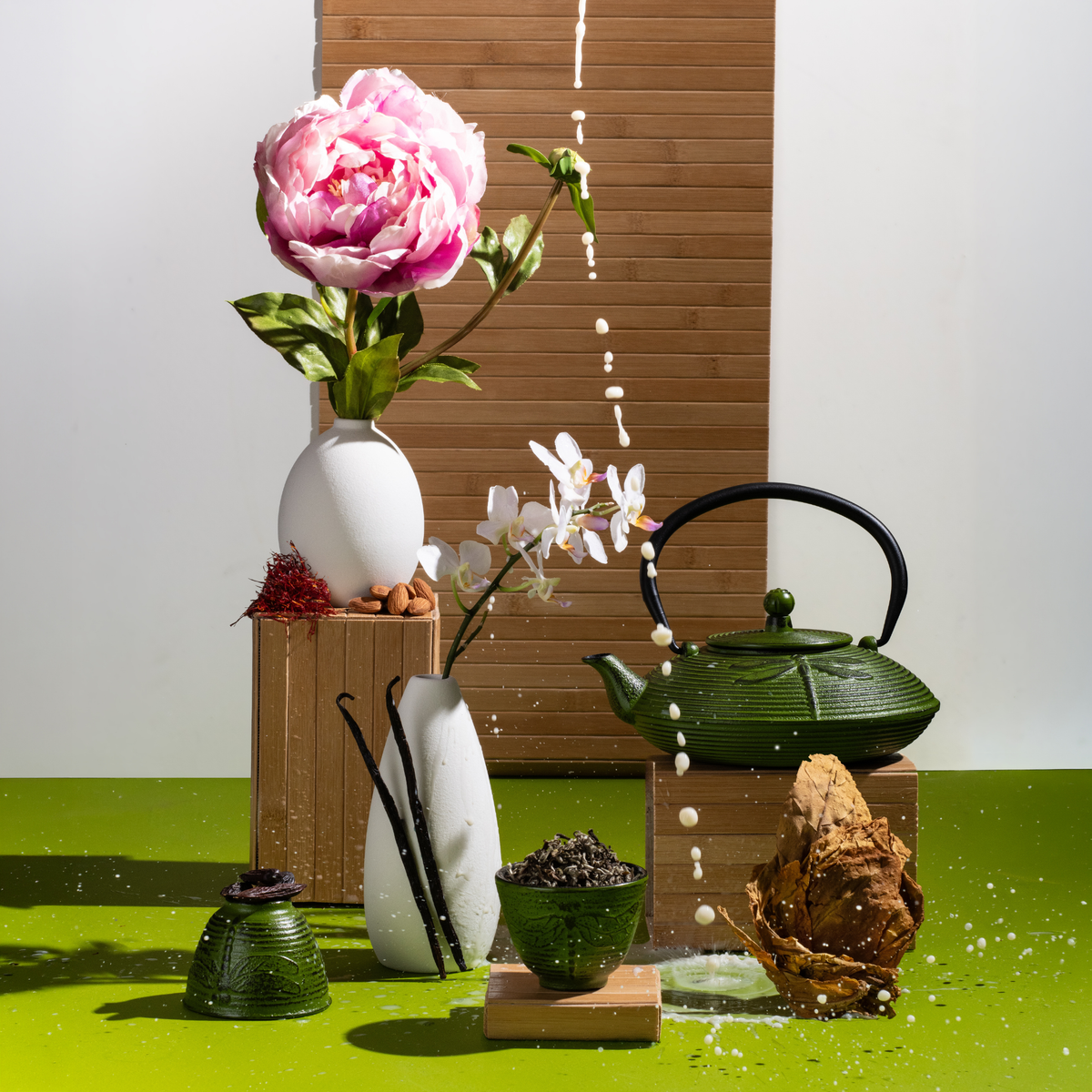 Styled scene with a pink peony in a white vase, a white orchid in a tall white vase, saffron threads and almonds, vanilla beans, a green cast-iron teapot and cup with loose tea, and dried tobacco leaves, all set against bamboo panels and a bright green surface. A stream of coconut milk falls from above, splashing droplets across the scene
