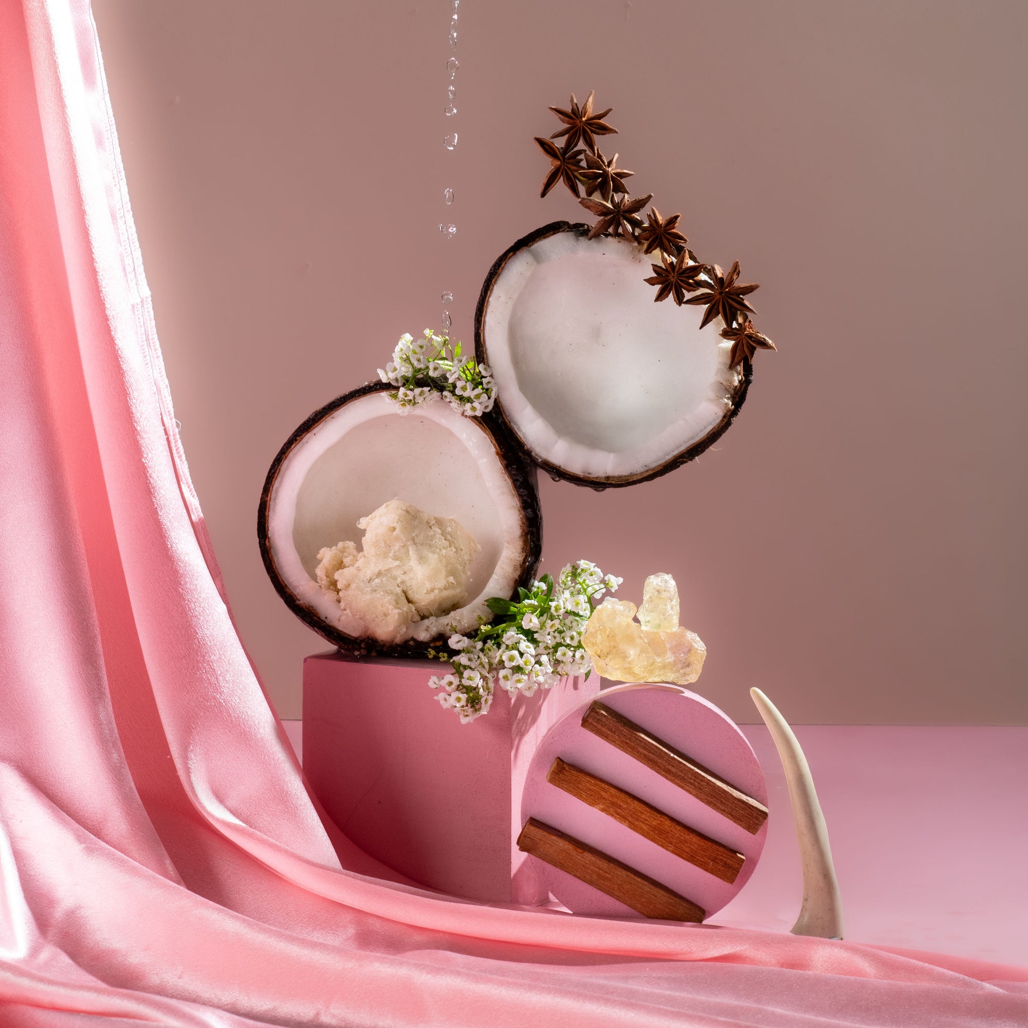 Decorative setup with coconut halves, flowers, and a pink container on a pink fabric background.