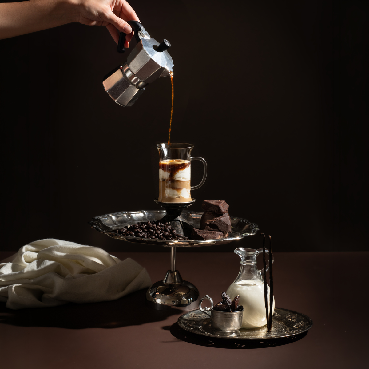 Moody still life on a dark brown background: a hand pours espresso from a stovetop moka pot into a glass mug layered with vanilla ice cream and milk. The mug sits on a silver pedestal tray with coffee beans and dark chocolate chunks, and a glass pitcher of cream, vanilla beans, and tonka beans sit on an ornate silver tray nearby.