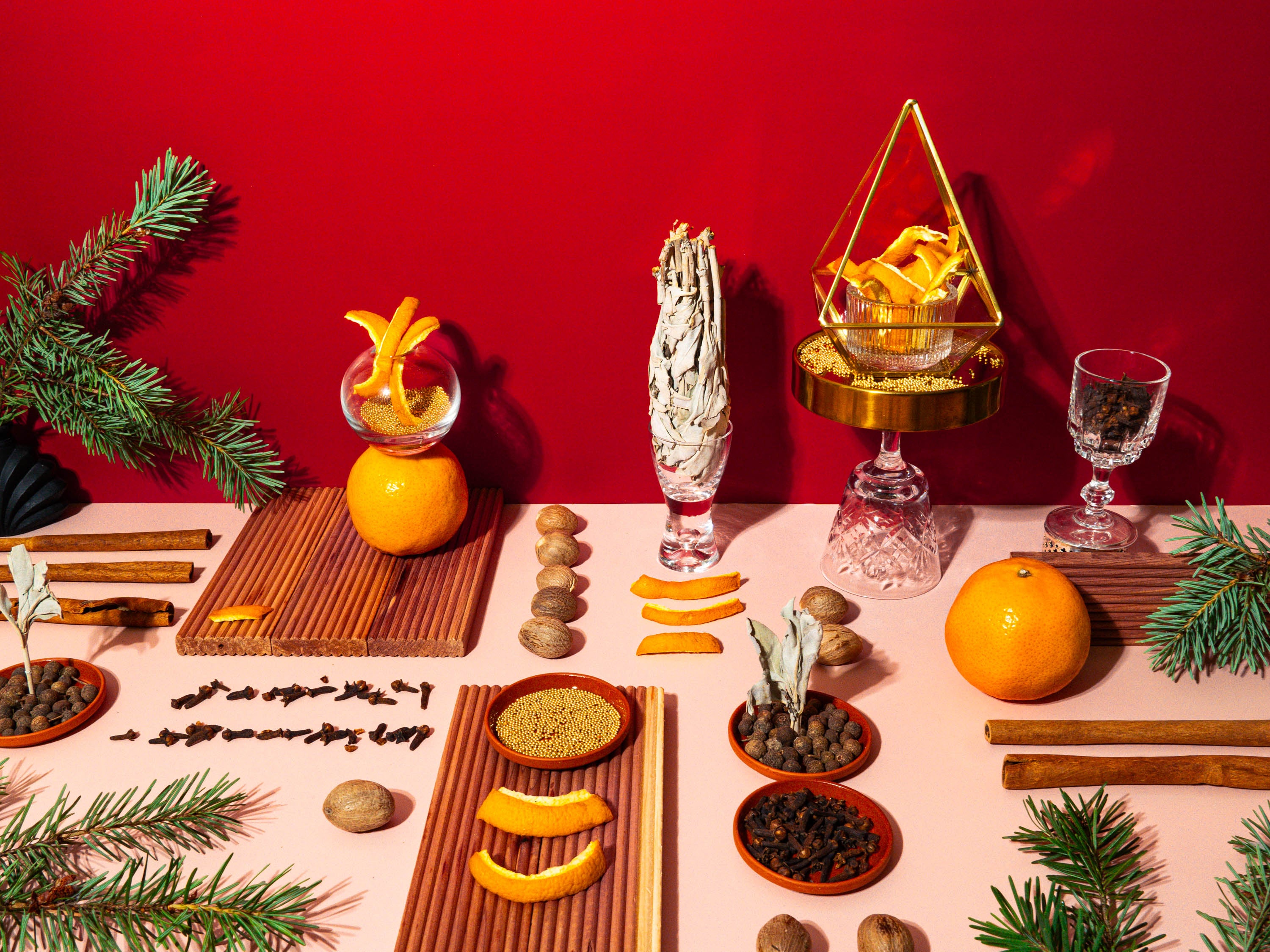 A vibrant holiday tablescape featuring an array of festive elements on a red background. Central to the image is a display of fresh and dried oranges, cinnamon sticks, cloves, and nutmeg, alongside evergreen branches and a pyramid-shaped terrarium filled with golden accents. On the left, an orange is adorned with a peel spiral standing on a wooden board, adding a touch of whimsy. Two elegant glassware pieces complete the scene, evoking a warm, celebratory mood reminiscent of Christmas spices and traditions