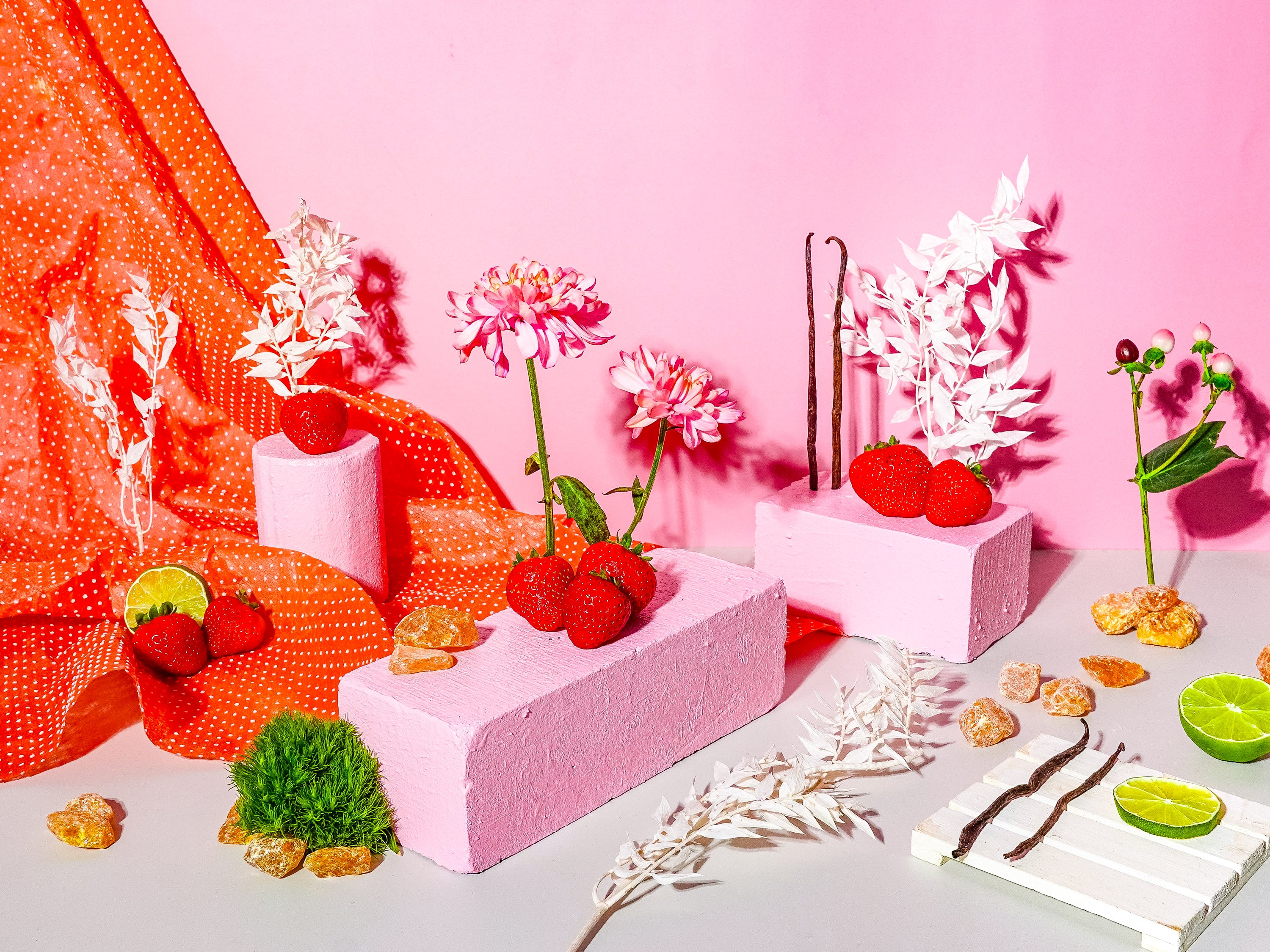 A bright, playful arrangement set against a pink backdrop with pops of red from strawberries and an orange polka-dotted fabric. Pink painted blocks hold fresh strawberries, vanilla pods, and delicate florals, while scattered lime slices, amber resin pieces, and greenery add freshness and texture. A whimsical and vibrant scene celebrating fruity and sweet elements.