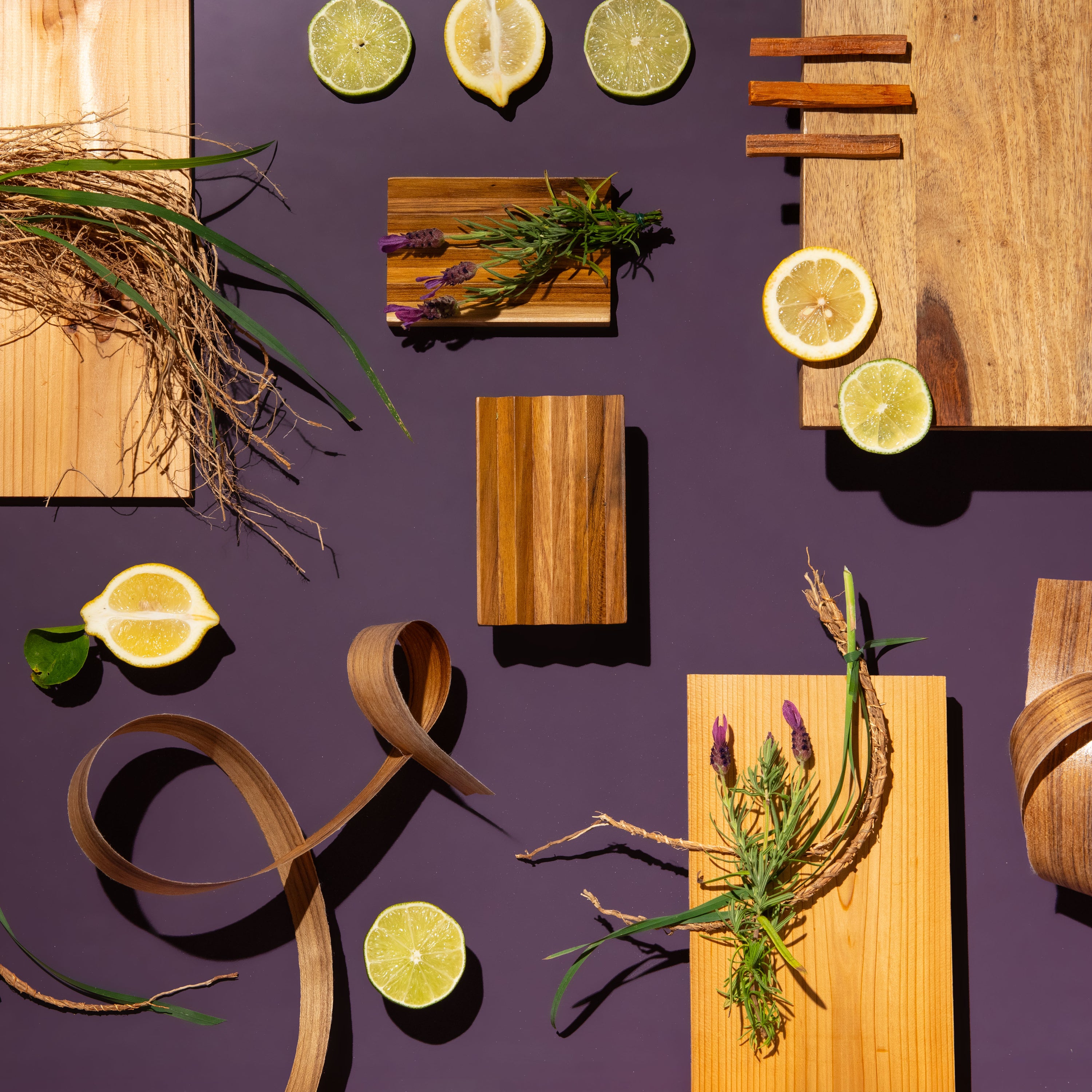 A styled flat-lay of multiple natural wood cutting boards in different shapes and tones, arranged on a dark purple background with decorative props including sliced lemons, cinnamon sticks, herbs, and dried grass.