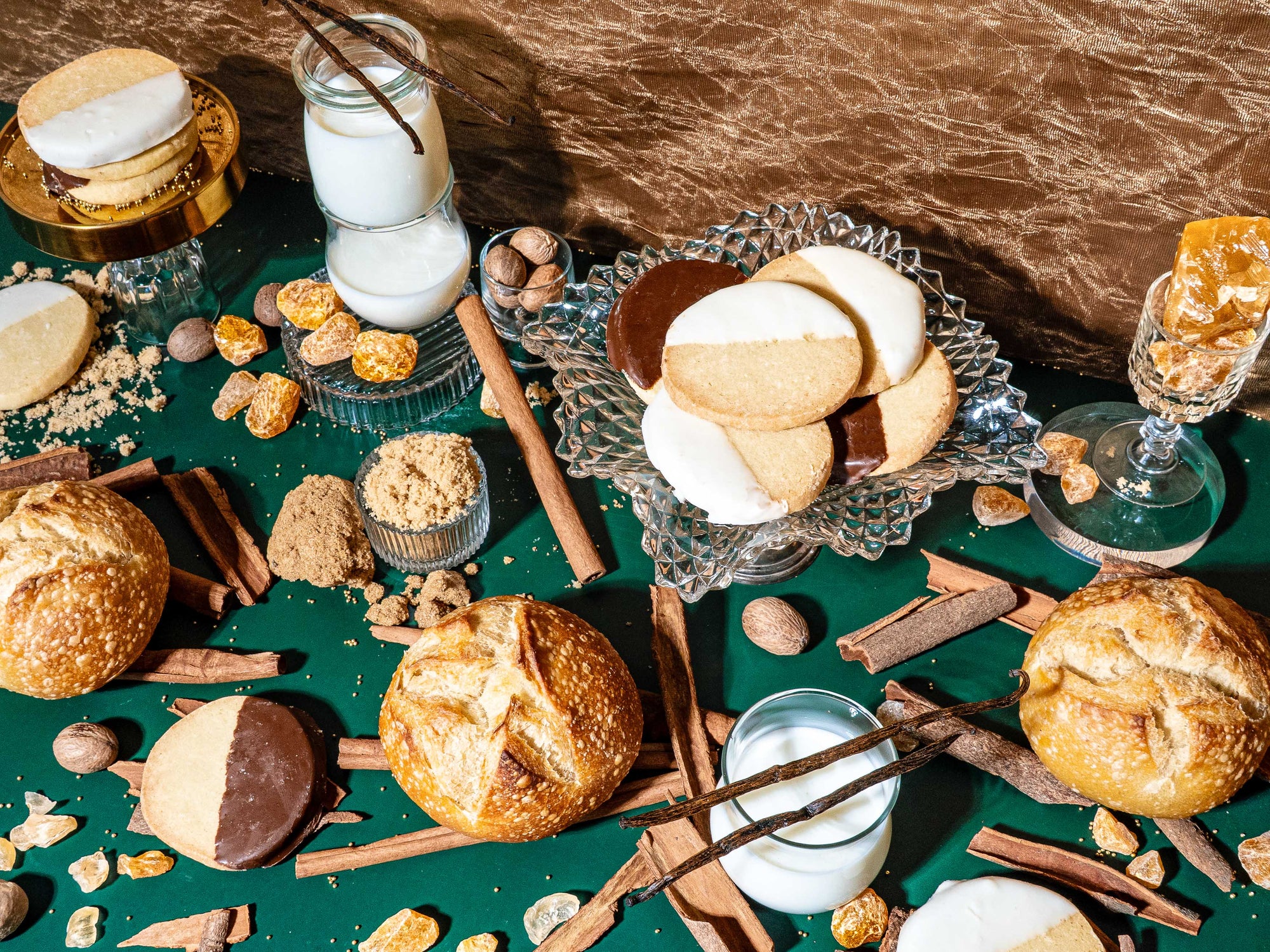 A festive, indulgent flat lay featuring black-and-white cookies, crusty bread rolls, glasses of milk, cinnamon sticks, vanilla beans, nutmeg, brown sugar, and shards of golden brittle. Set against a deep green tablecloth and gold textured backdrop, the scene evokes cozy holiday baking with a rich, nostalgic warmth.