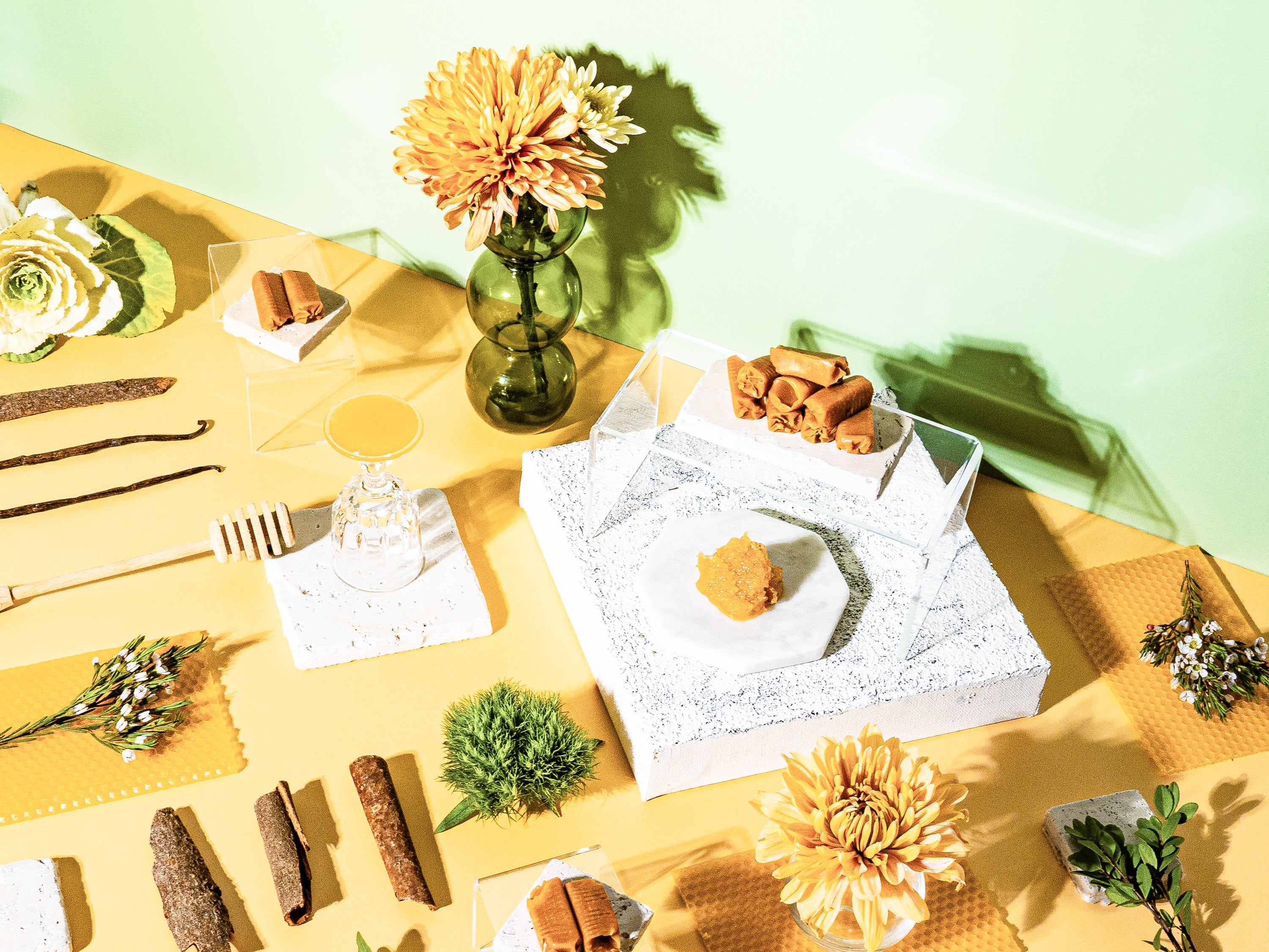 A vibrant flat-lay featuring honeycomb, cinnamon sticks, vanilla beans, and a selection of natural elements like flowers, greenery, and wax materials. The composition evokes warmth and sweetness, with a modern aesthetic showcasing honey drizzler, floral accents, and a soft yellow and green backdrop. A perfect representation of nature-inspired fragrances and cozy indulgence.