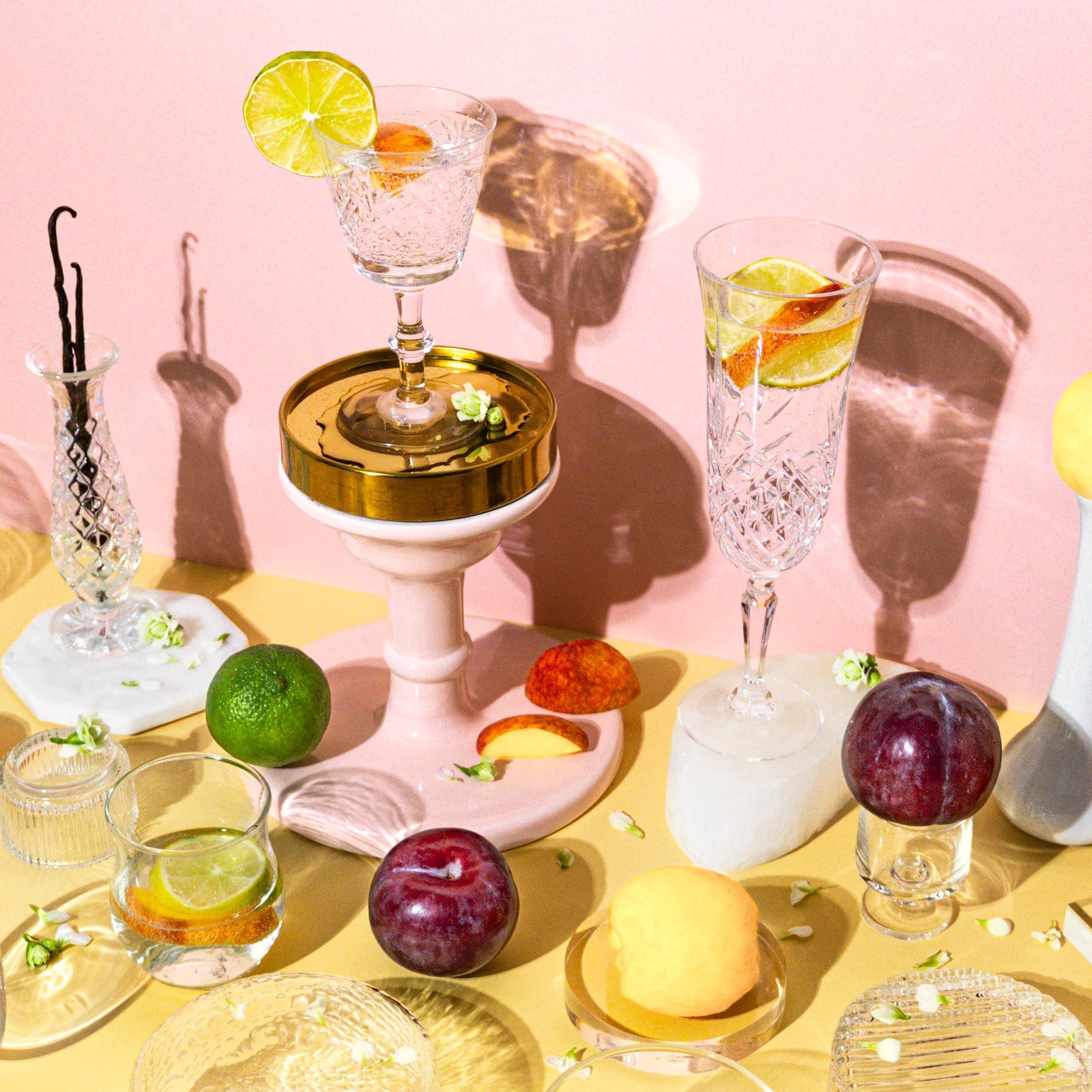 An assortment of fragrance oils presented in small glass bottles, surrounded by an arrangement of fruits including peach, lime, and plum, with a backdrop featuring a drink setup with cocktails and vanilla beans.